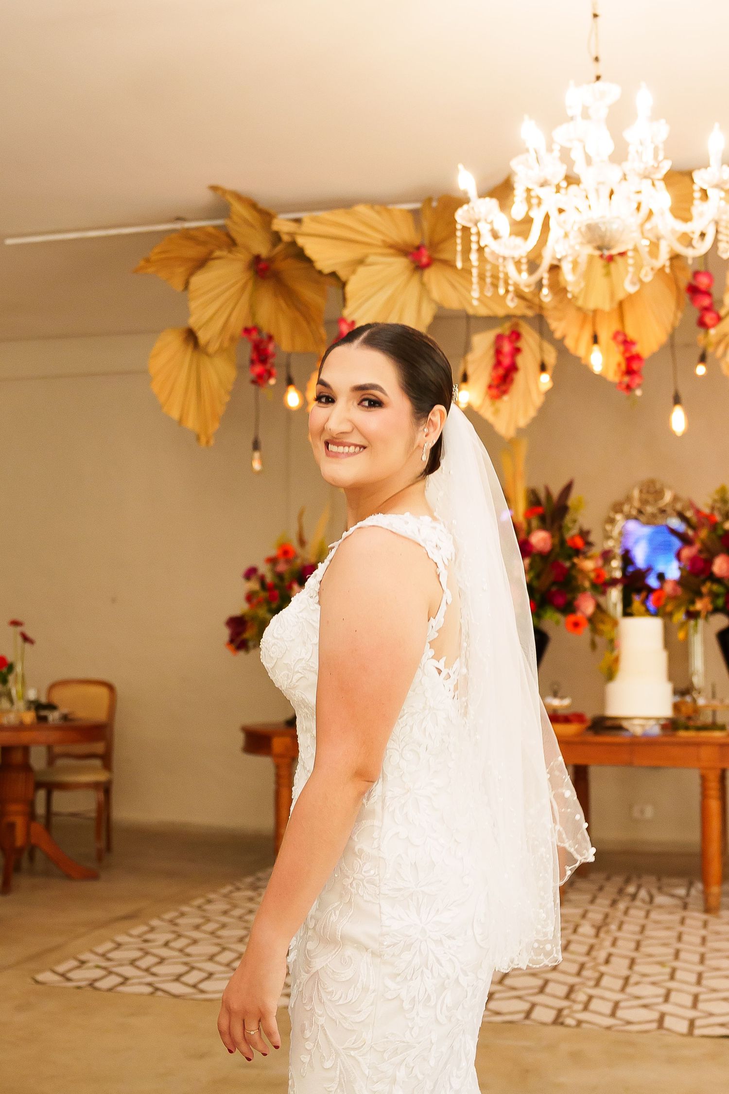 Smiling bride in white lace gown and veil poses in decorated venue with chandelier and colorful floral backdrop.