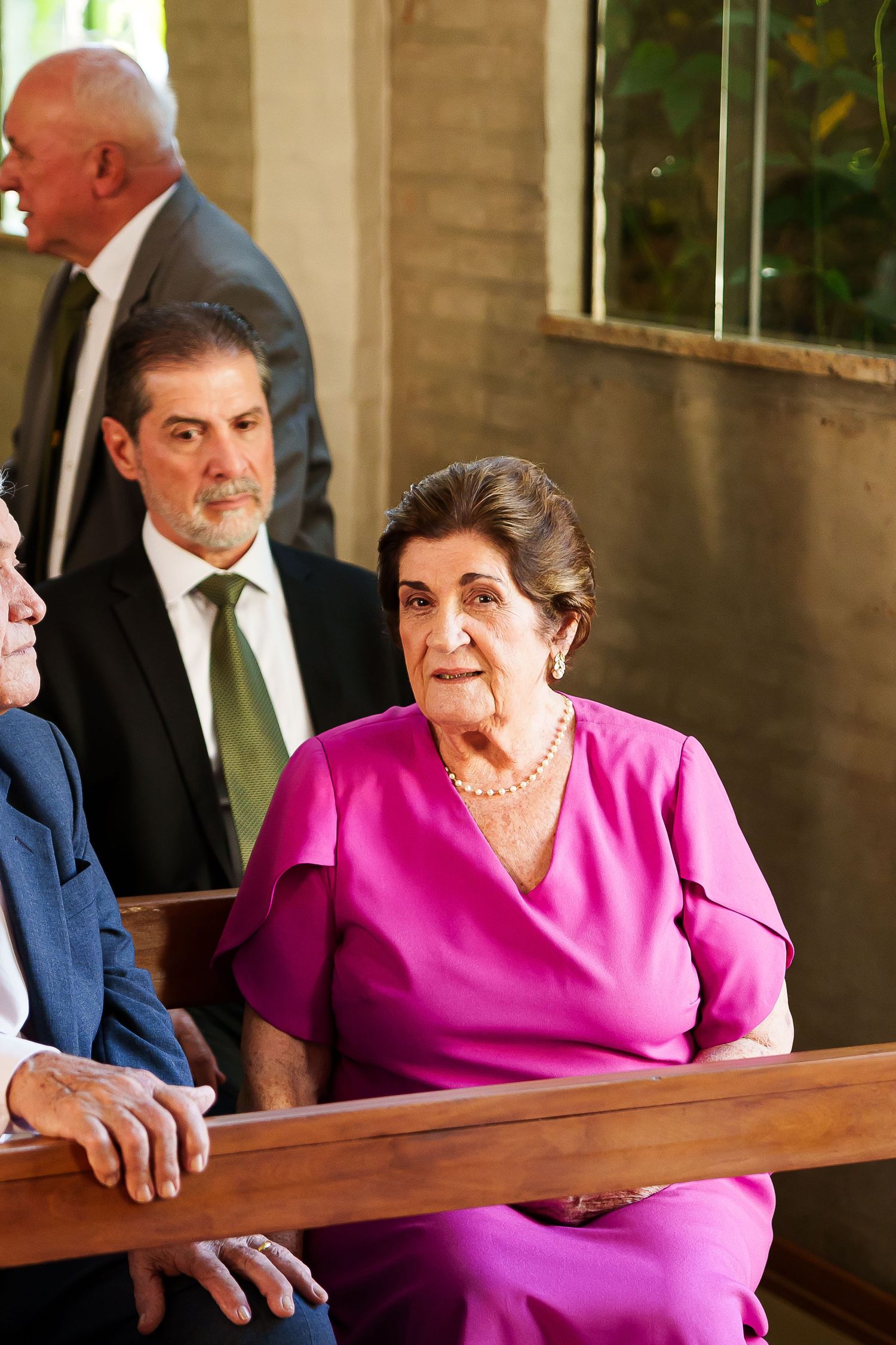 Elderly woman in pink dress seated on church pew at formal event with suited guests in background.