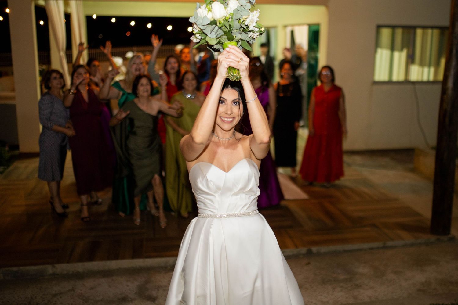 Bride in white gown tosses bouquet to excited guests at wedding reception.