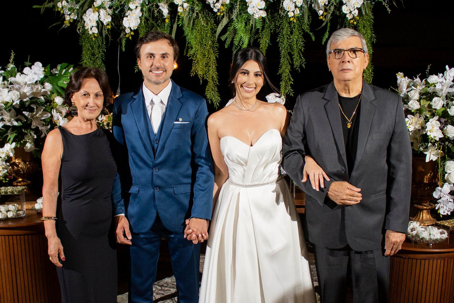 Wedding couple in formal attire poses with two guests before floral backdrop at elegant ceremony venue.