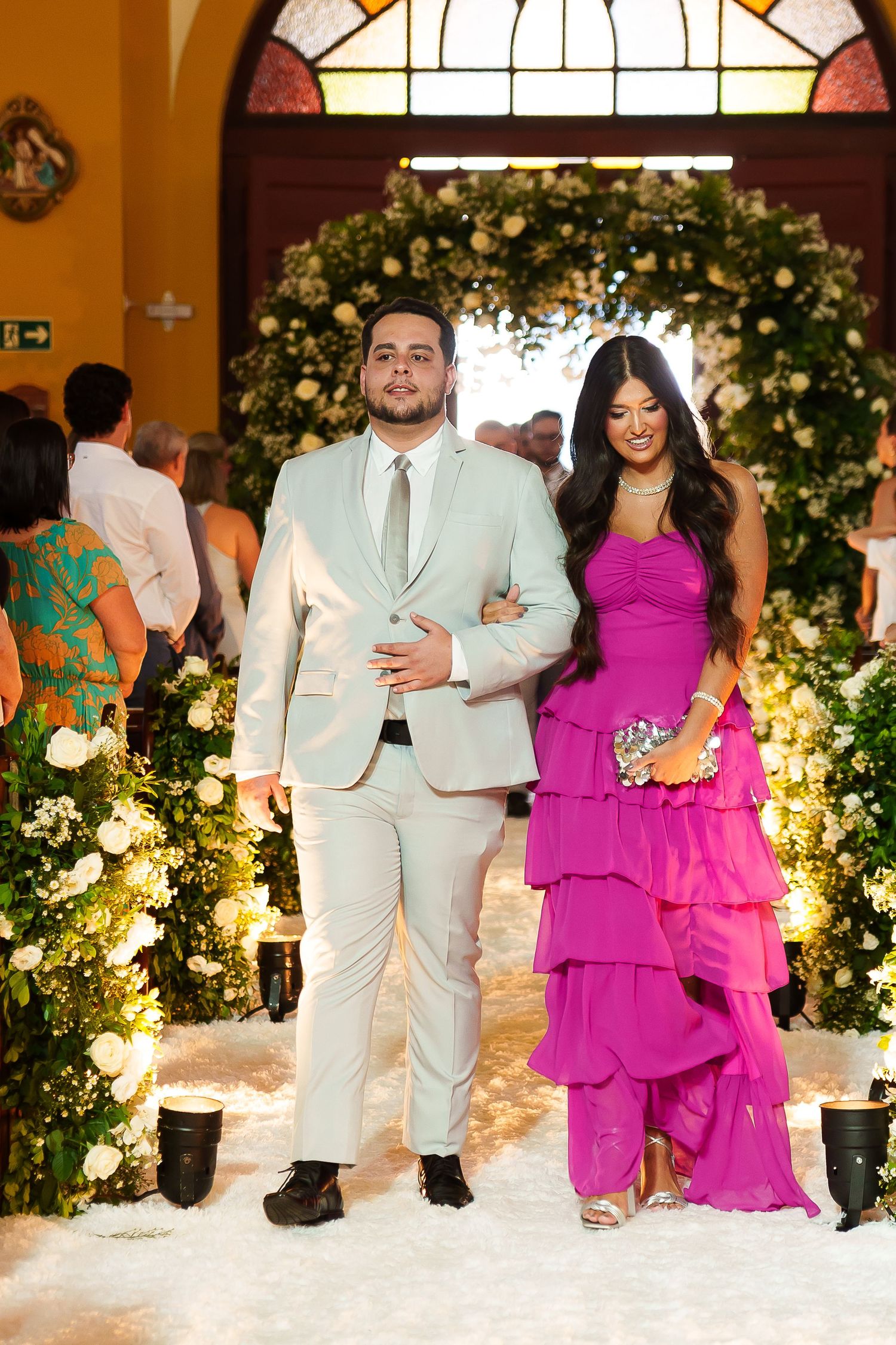 Couple walking down floral wedding aisle, man in cream suit, woman in tiered magenta gown, elegant church setting.