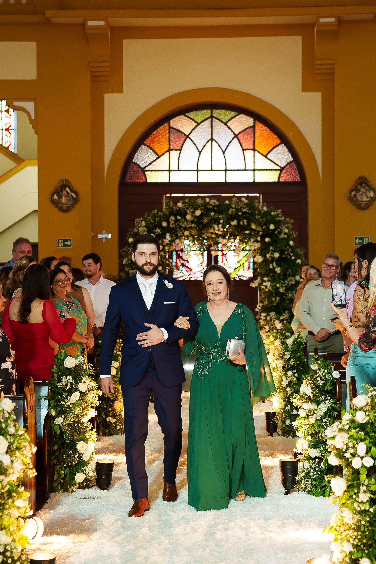 Groom in navy suit escorts woman in emerald green gown down floral-lined church aisle during wedding ceremony.