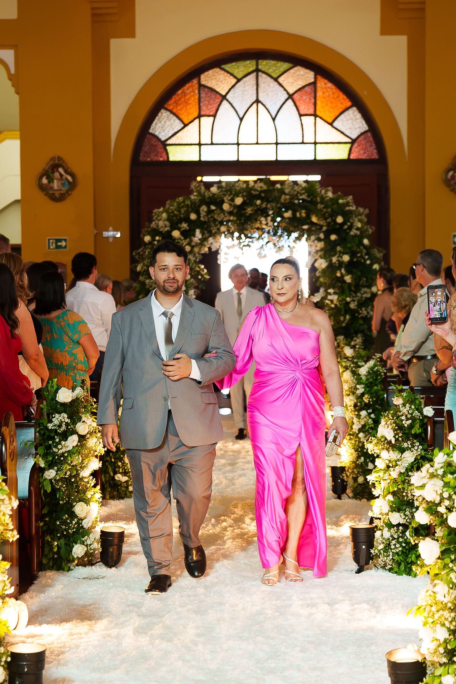 Couple walks down church aisle, man in gray suit, woman in pink one-shoulder gown, floral arch backdrop.