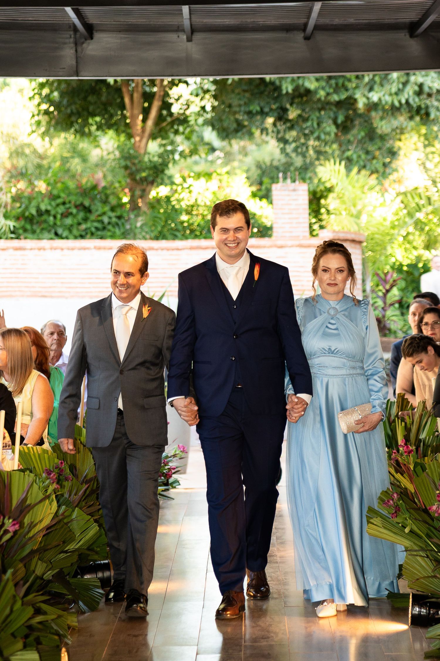 Groom walks down the aisle with parents at an outdoor wedding ceremony surrounded by tropical greenery.