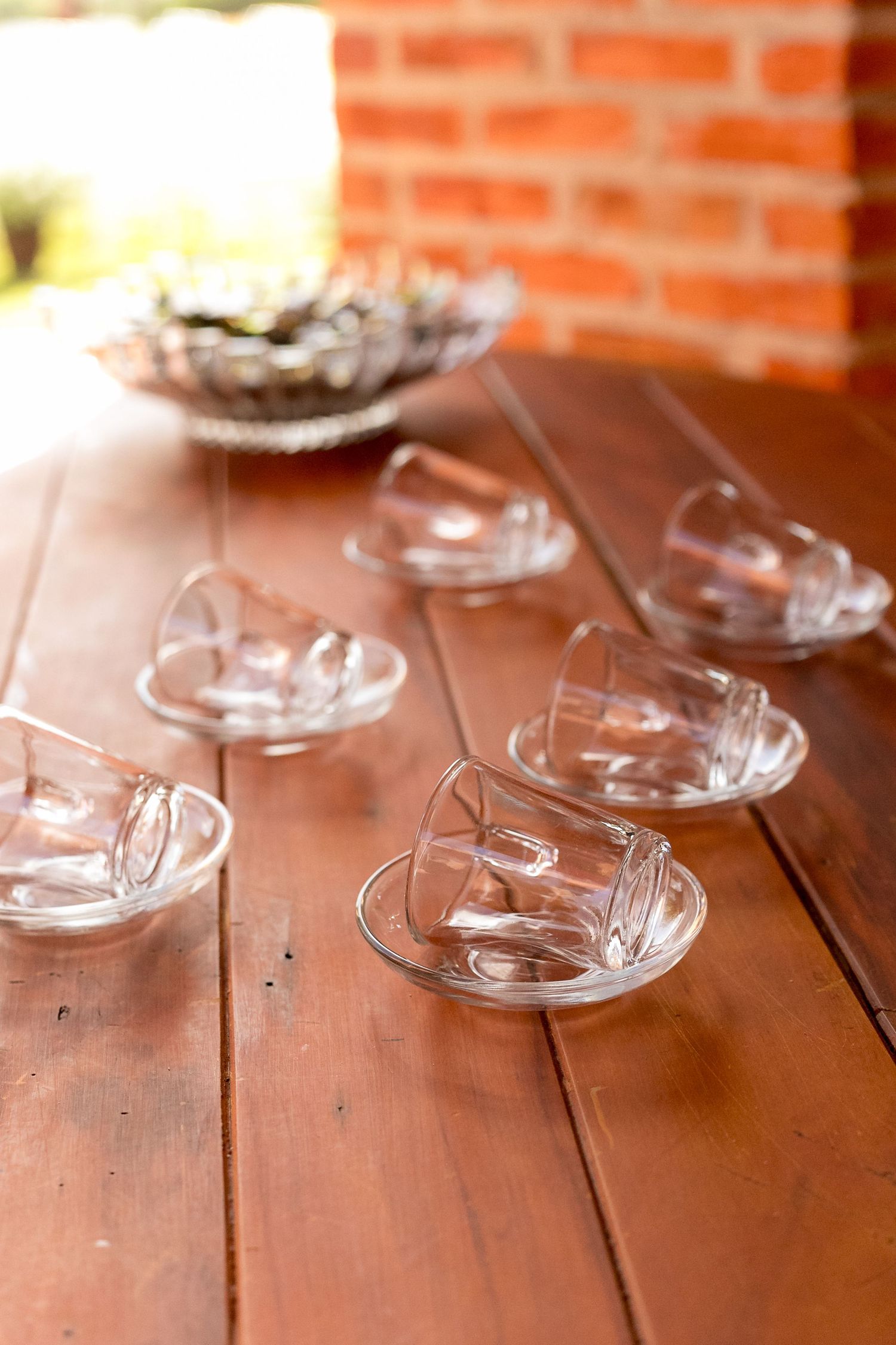 Clear glass teacups and saucers arranged on a wooden table with a brick wall background.