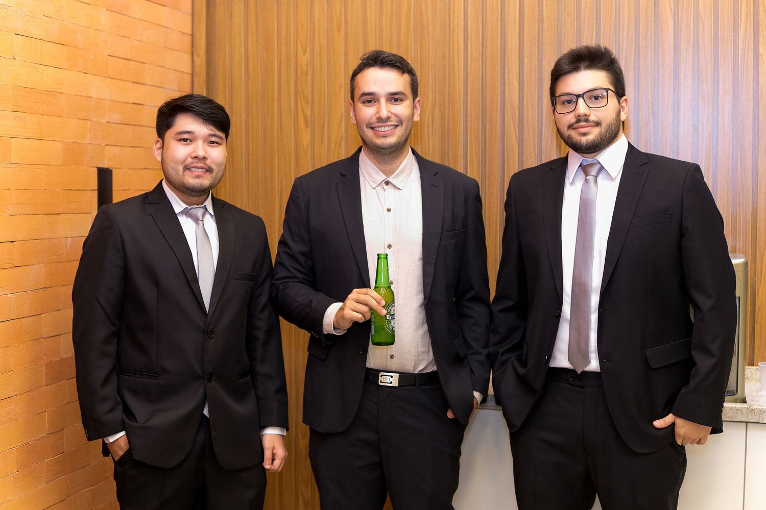 Three young men in dark suits smiling together at a formal event, one holding a green bottle.