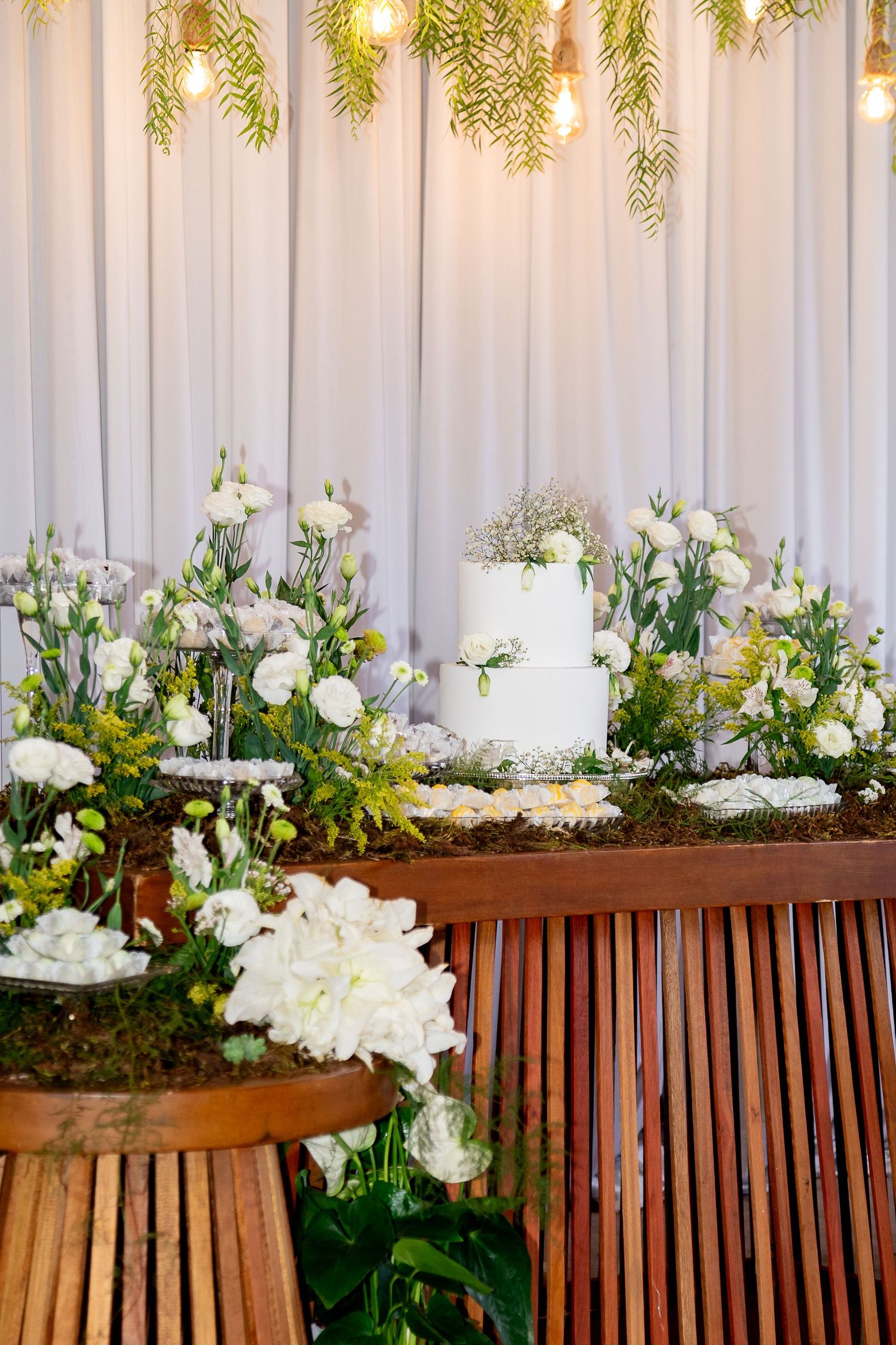 Elegant wedding cake display with white flowers, greenery, and warm Edison bulb lights on a rustic wooden table.