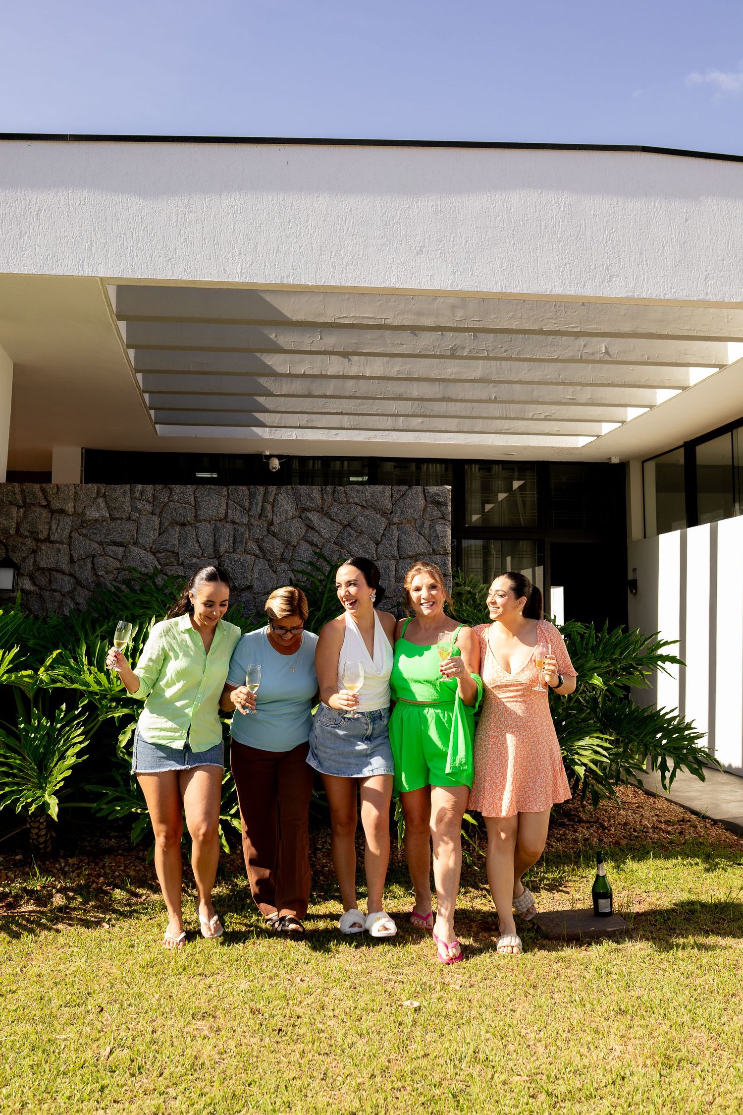 Five women smiling and holding champagne glasses outdoors in front of a modern house on a sunny day.