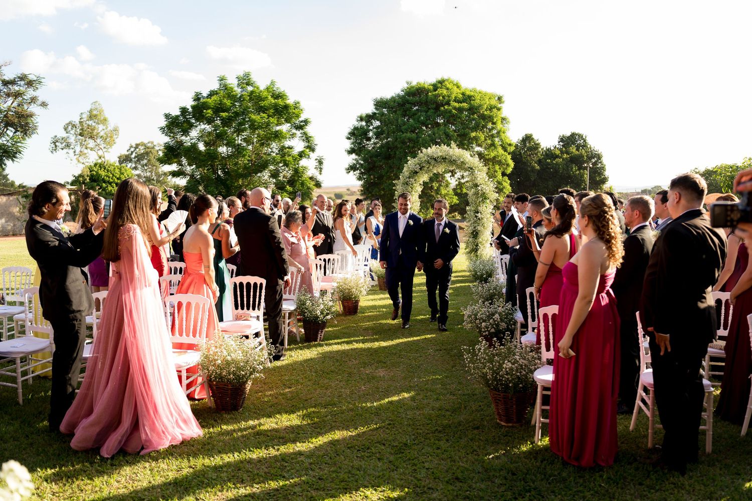 Outdoor wedding ceremony on green lawn with guests standing, bridal party in pink and red gowns, floral arch in background.