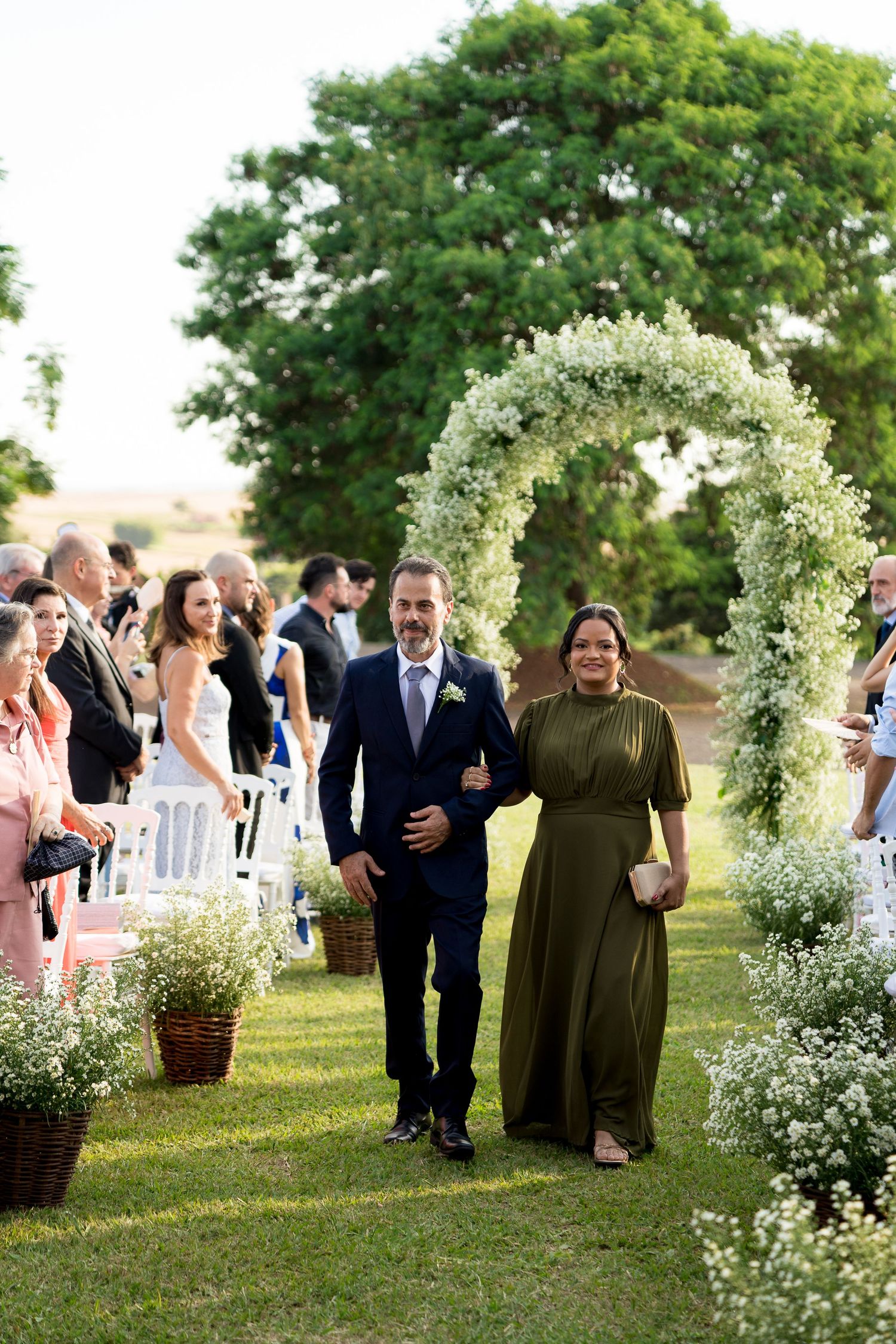 Couple walks down outdoor wedding aisle under white floral arch, guests seated on white chairs, lush green trees in background.