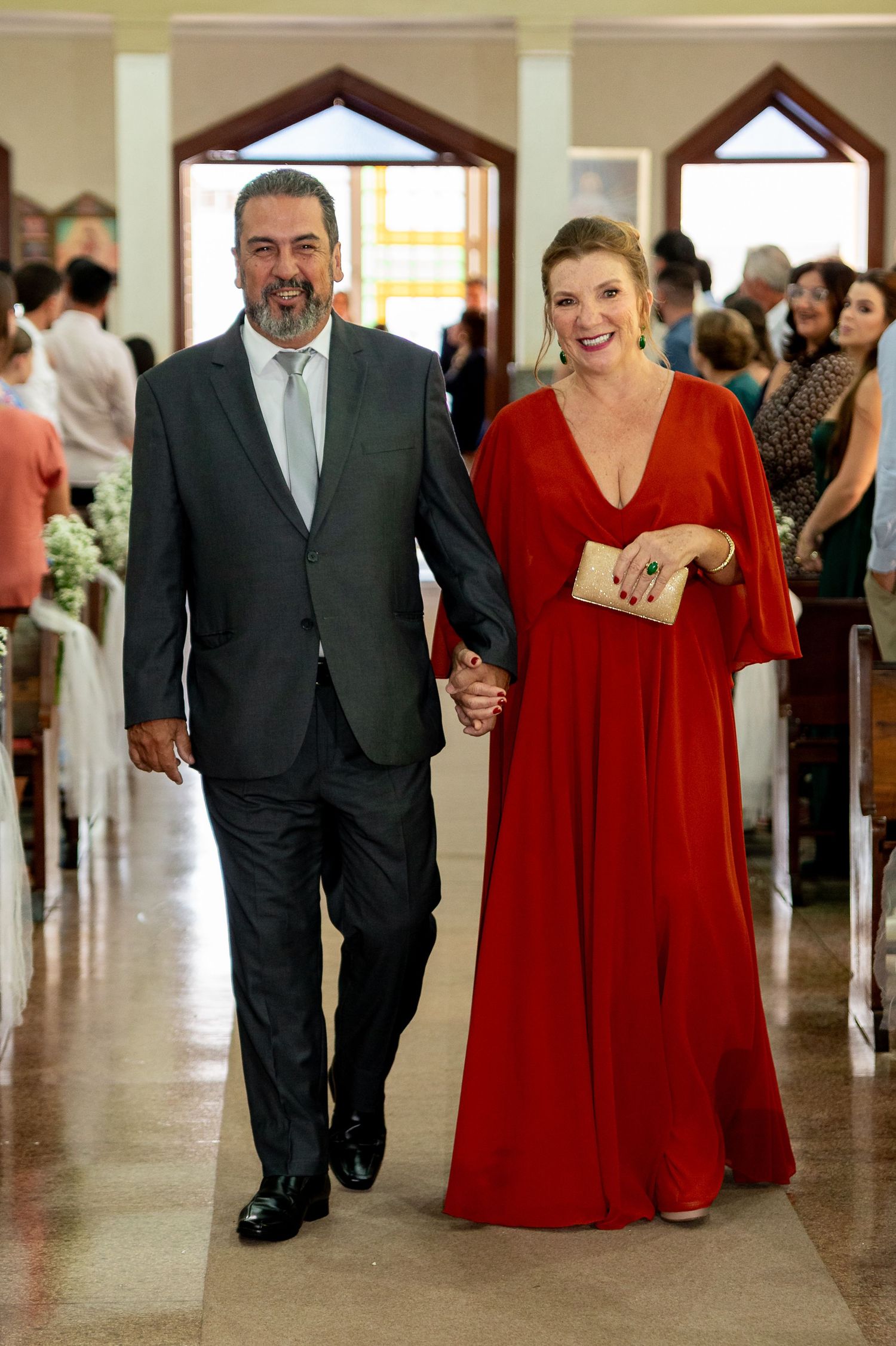 Elegant couple walking down church aisle, man in gray suit, woman in red gown, holding hands at wedding ceremony.