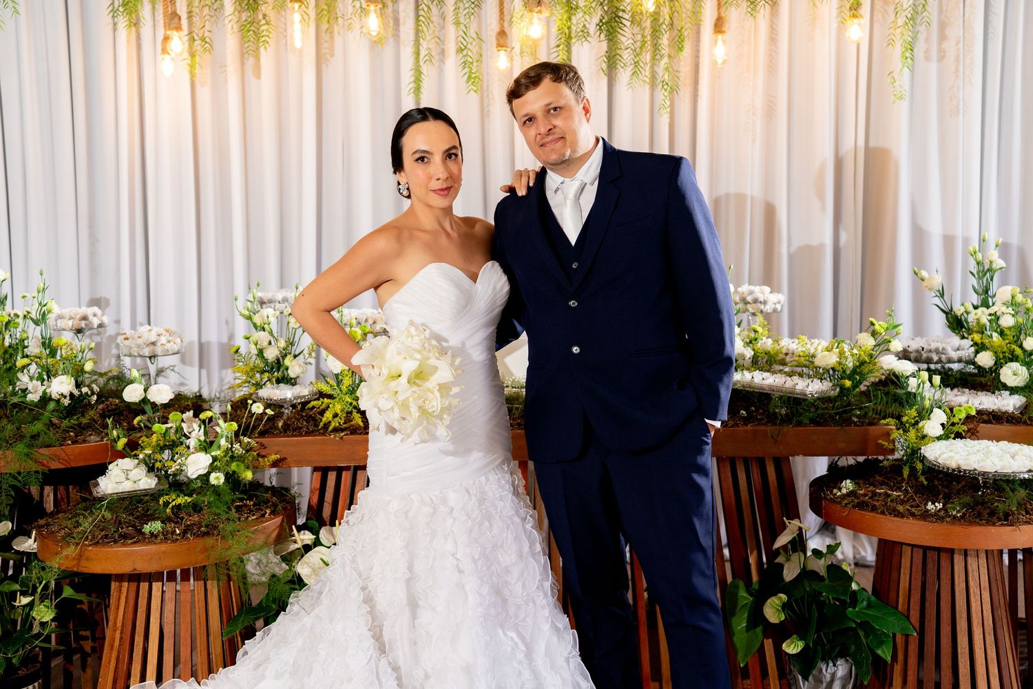 Bride in mermaid gown and groom in navy suit pose together at elegant floral wedding ceremony backdrop.