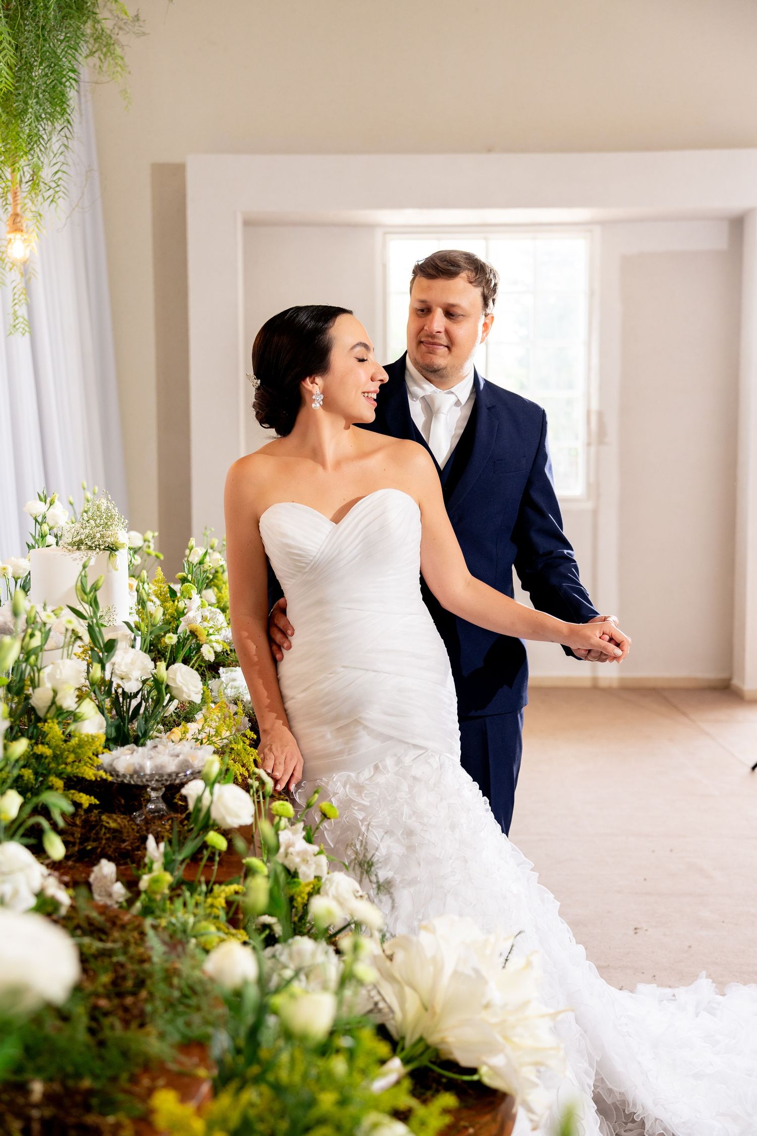 Bride in white mermaid gown and groom in navy suit hold hands near floral arrangements at elegant wedding venue.