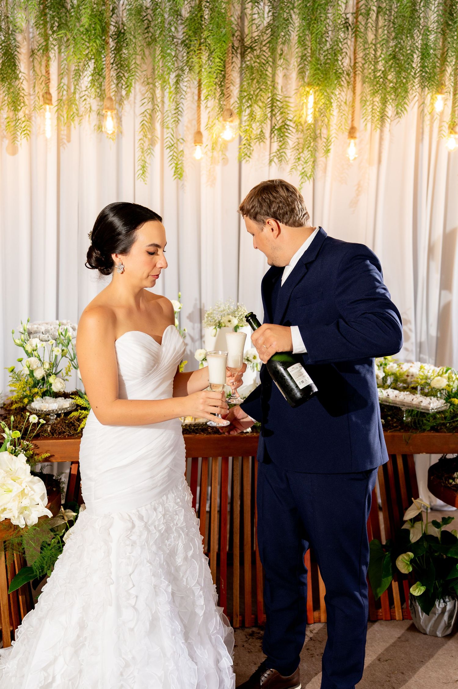 Bride and groom pouring champagne at wedding reception with floral backdrop and hanging Edison bulb lights.
