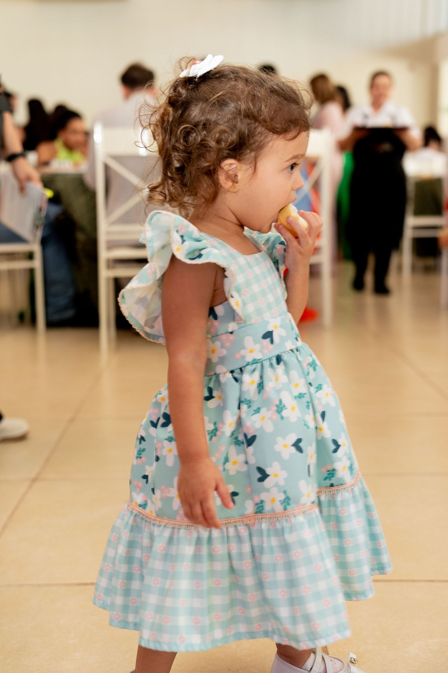 Toddler girl in teal floral gingham dress eating a cookie at an indoor party event.
