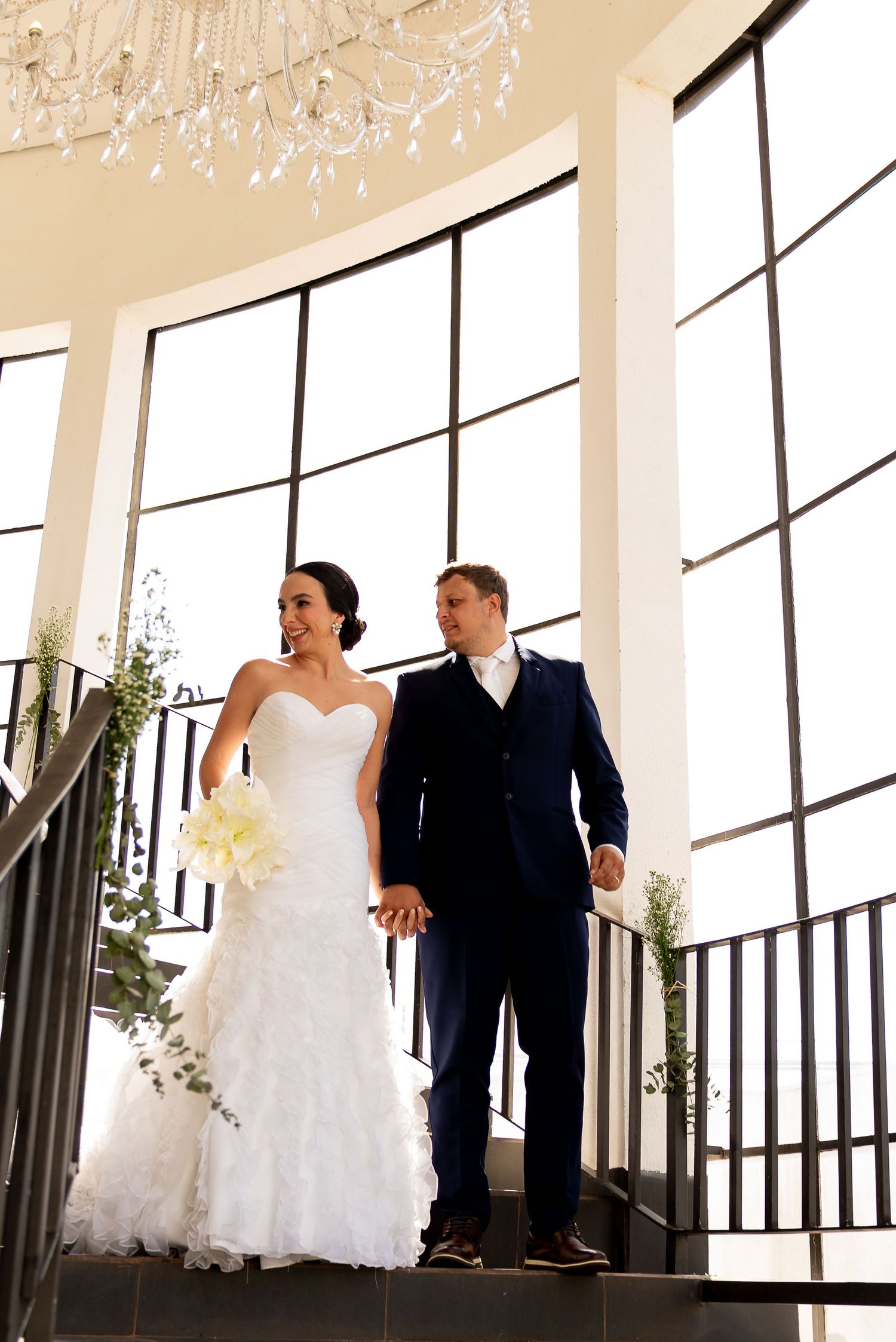 Bride and groom holding hands on elegant staircase with chandelier and large windows in a bright wedding venue.