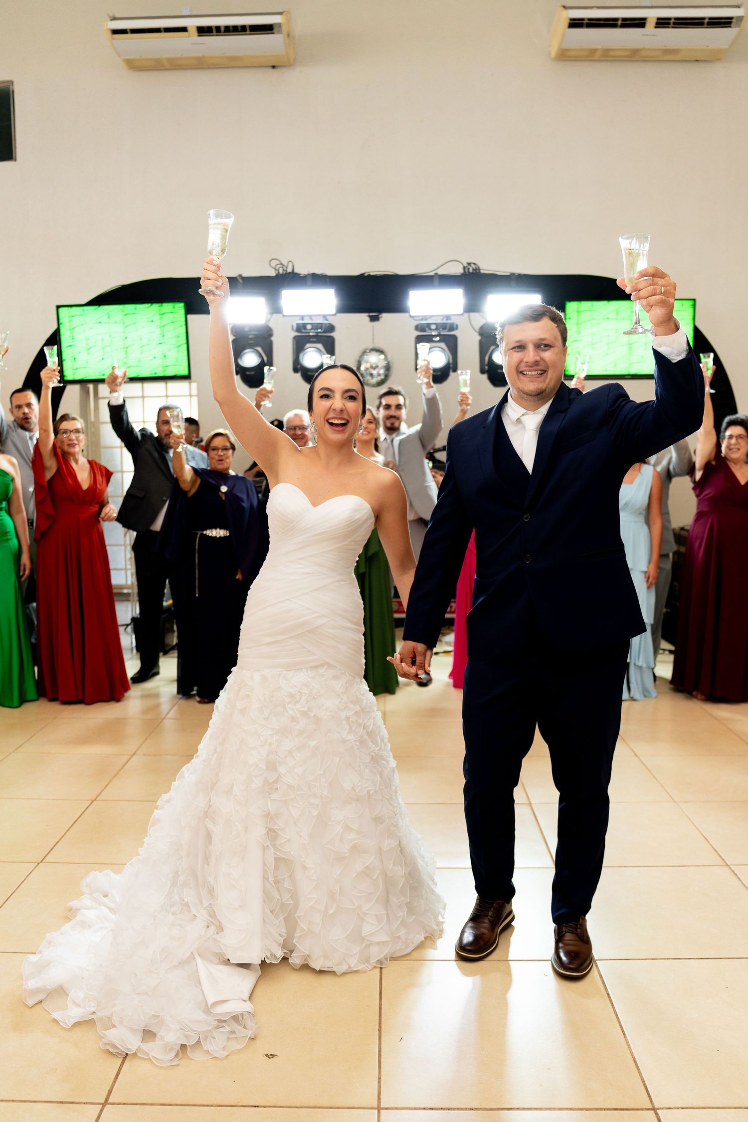 Bride and groom raising champagne glasses at wedding reception with guests celebrating in background.