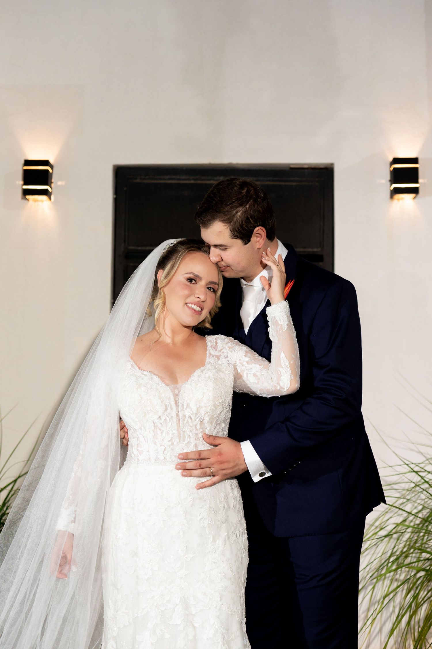 Bride in lace gown and veil smiles as groom in navy suit kisses her cheek at their wedding.