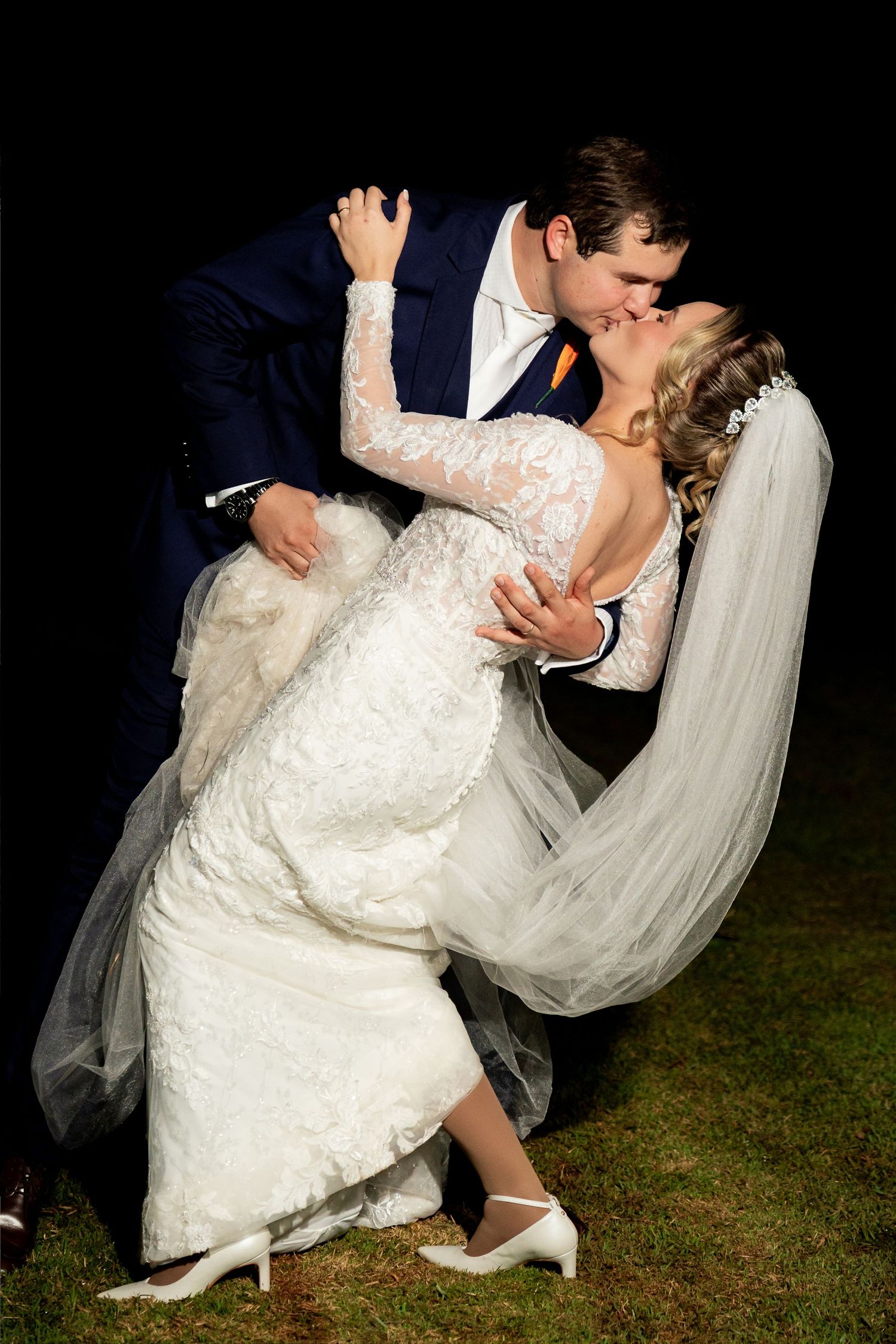 Bride and groom share a romantic dip kiss outdoors at night, bride in lace gown with veil, groom in navy suit.