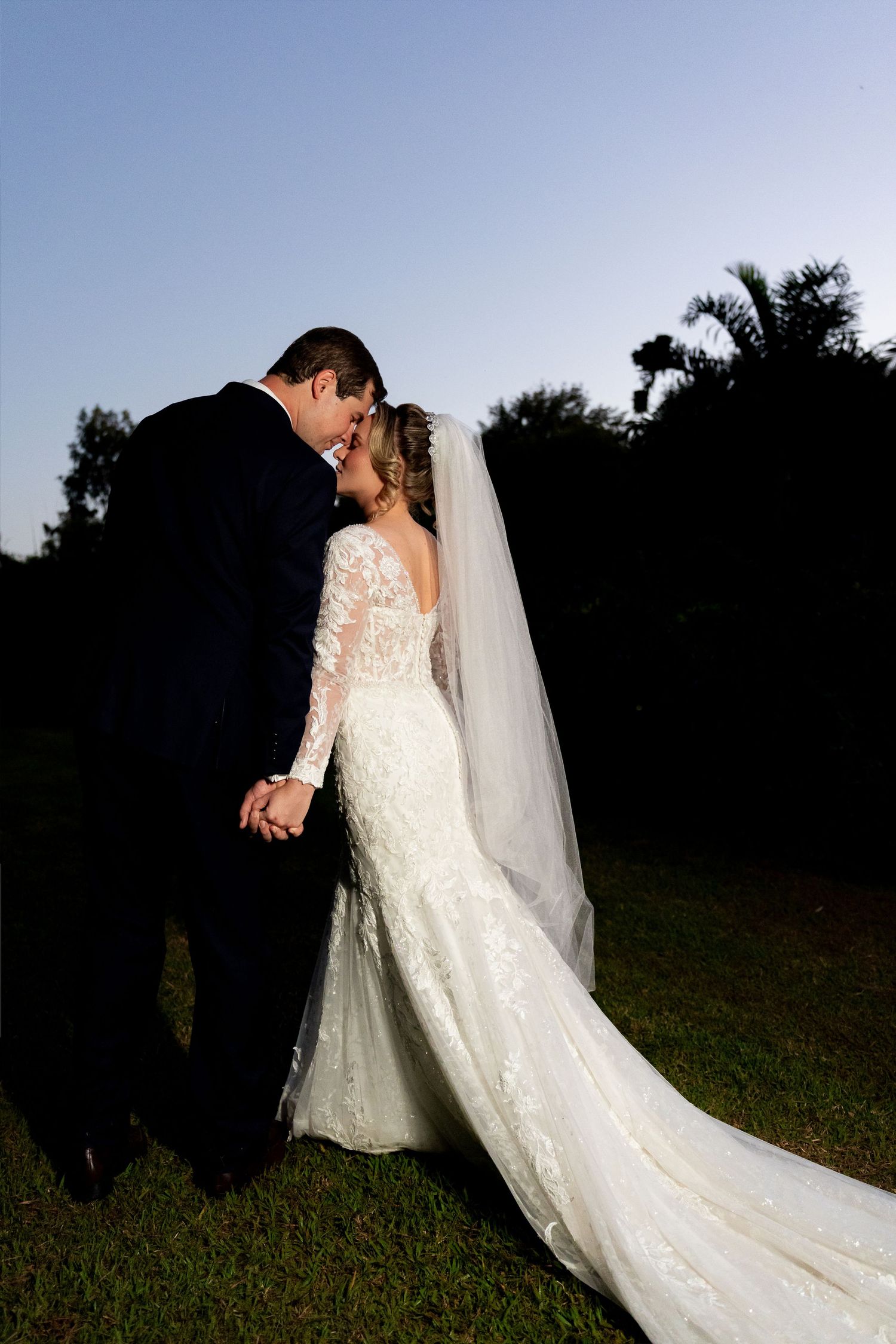 Bride and groom share a romantic forehead touch at dusk, her lace gown and veil trailing on lush green grass.