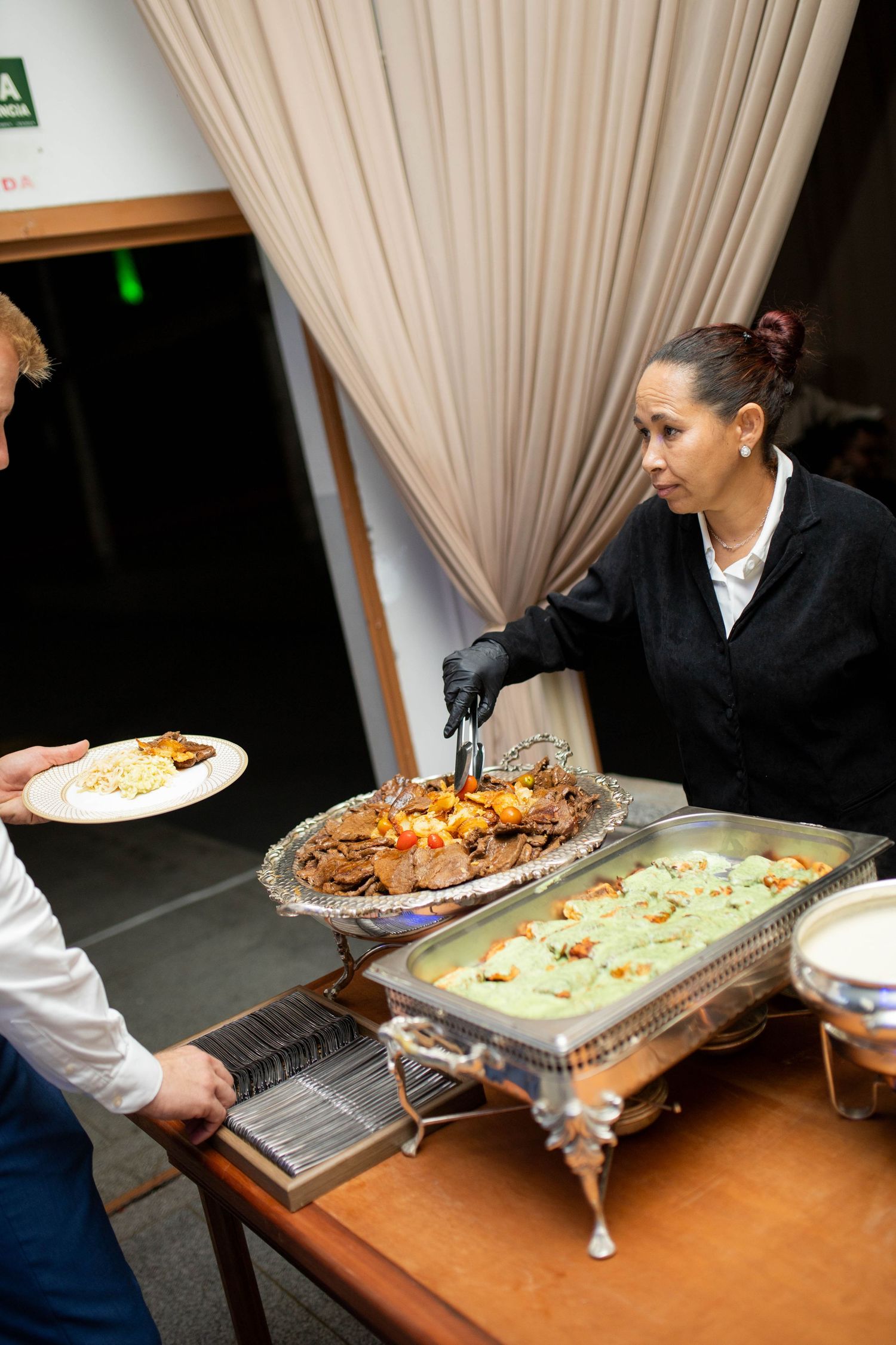 Catering staff serving food from chafing dishes at a buffet event, wearing black gloves and uniform.
