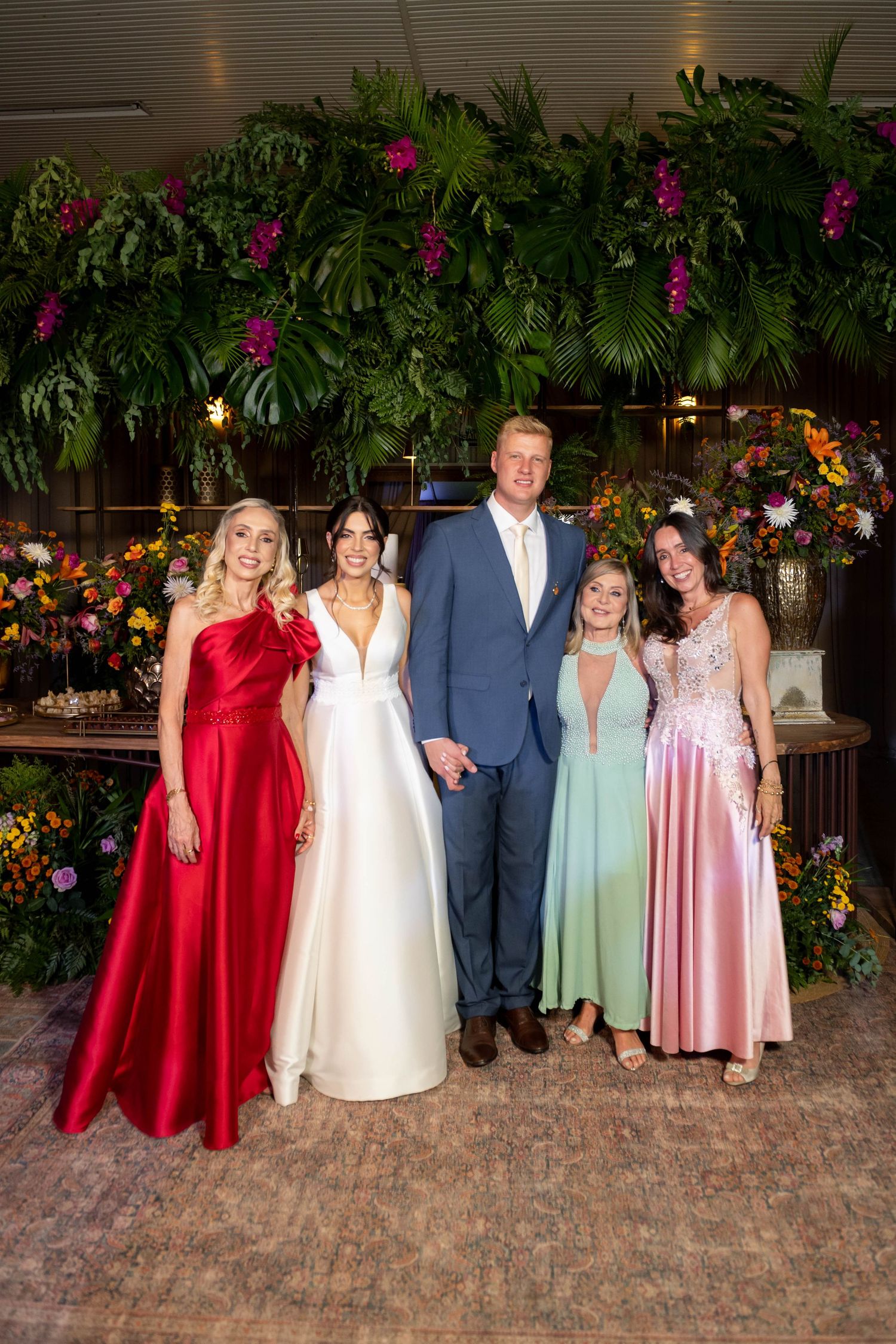 Wedding party poses before lush tropical floral backdrop, bride in white gown with groom and guests in elegant formal attire.