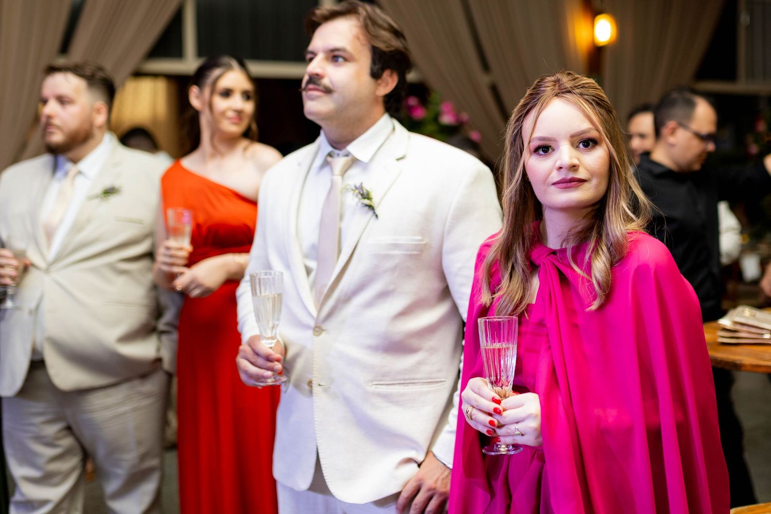 Elegantly dressed couple at a wedding reception, man in white suit with mustache, woman in pink cape dress holding champagne glasses.