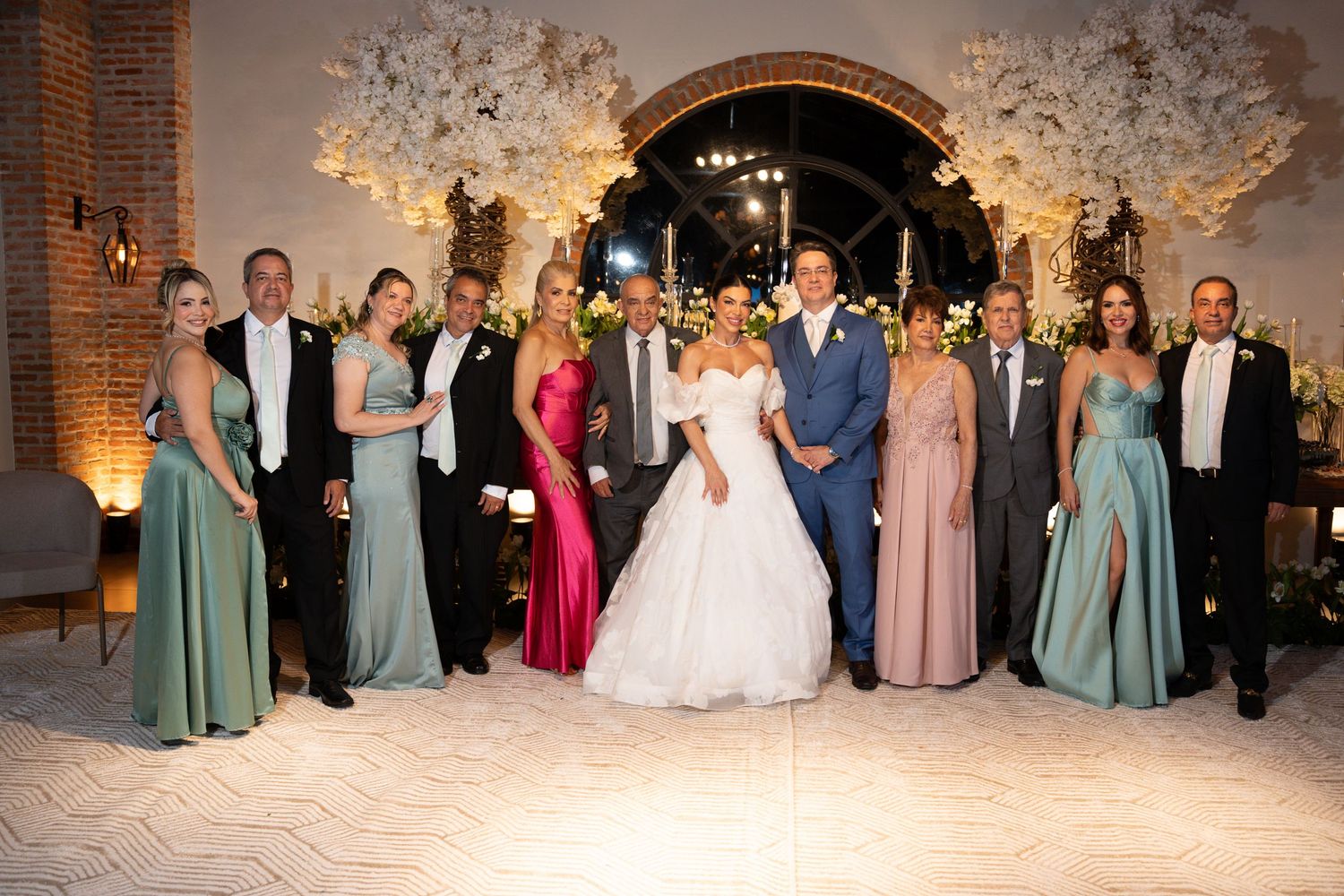 Wedding party poses together in elegant venue with floral arch, bride in white gown, bridesmaids in green and pink dresses.