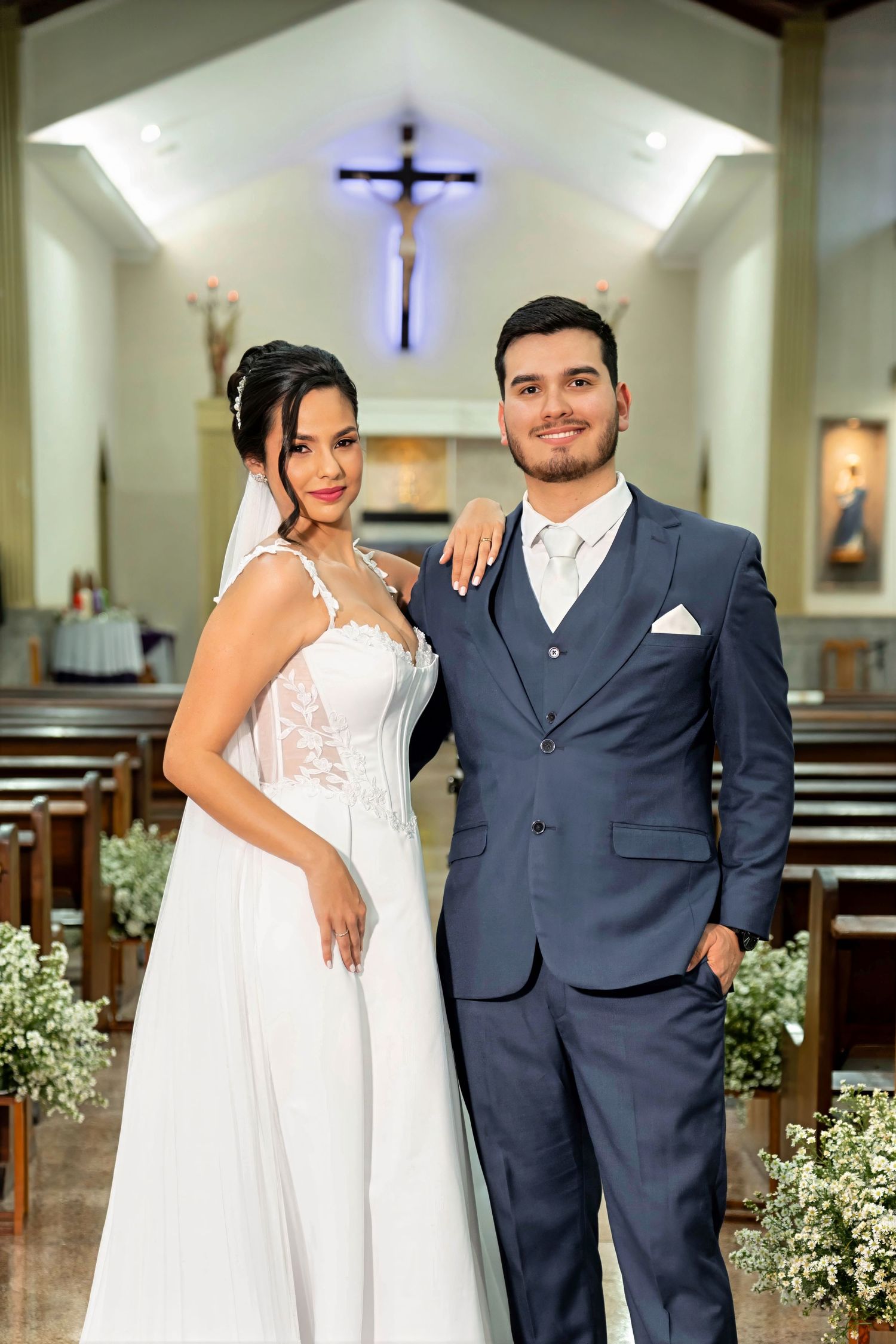 Bride in white lace gown and groom in blue suit pose together inside a decorated Catholic church on their wedding day.