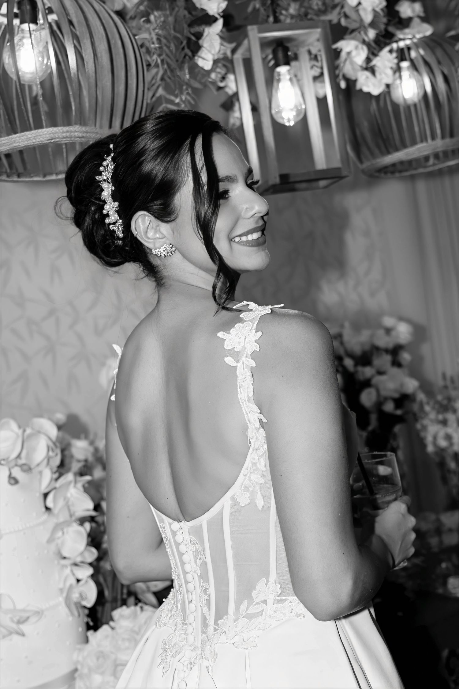 Black and white photo of a smiling bride in a backless lace wedding gown with floral hair accessory near a wedding cake.