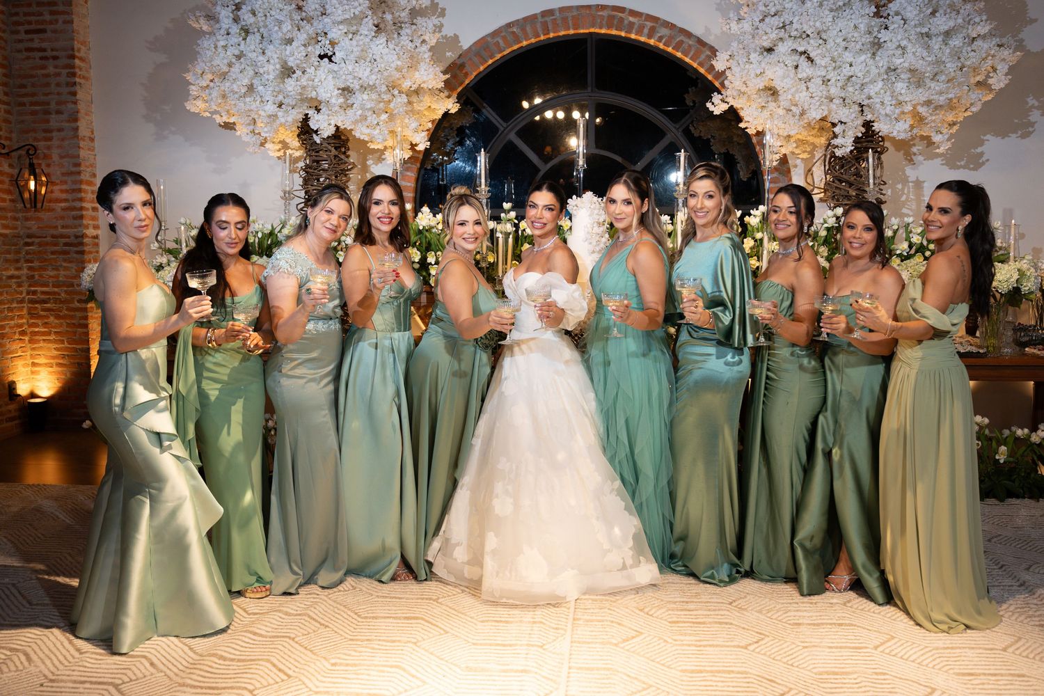 Bride in white gown poses with bridesmaids in sage green dresses at elegant floral wedding venue.