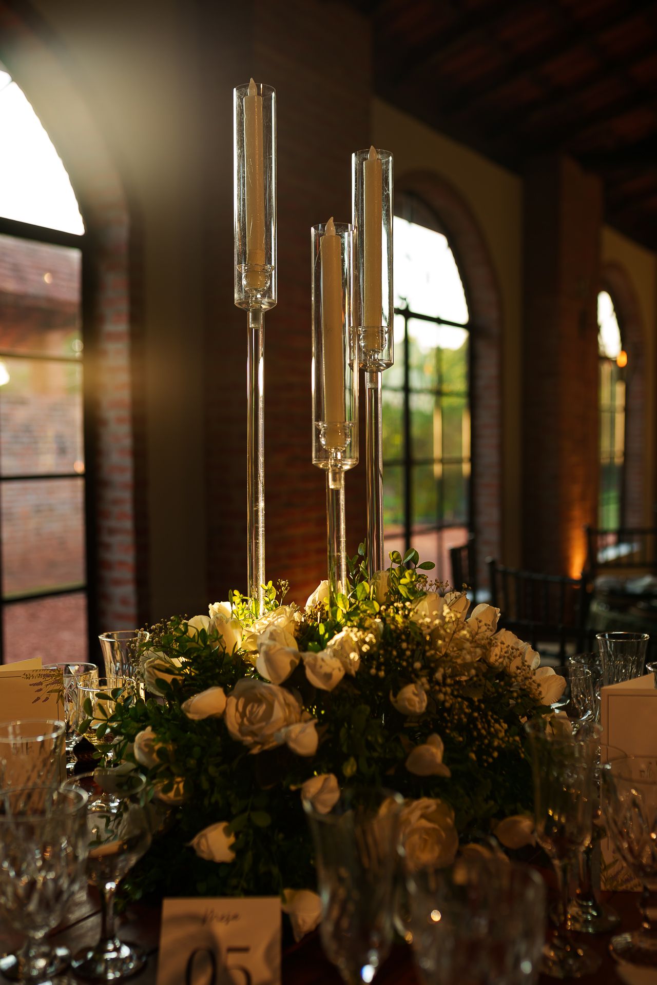 Elegant wedding centerpiece with tall glass candelabras and white roses on a reception table in a warmly lit venue.