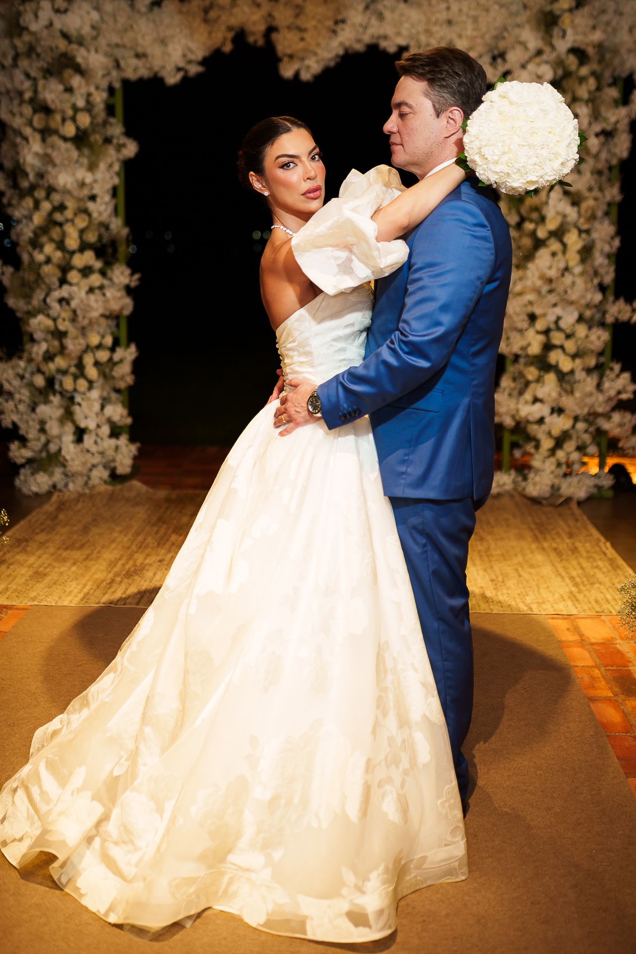 Bride in white ball gown and groom in blue suit share first dance surrounded by white floral arrangements.
