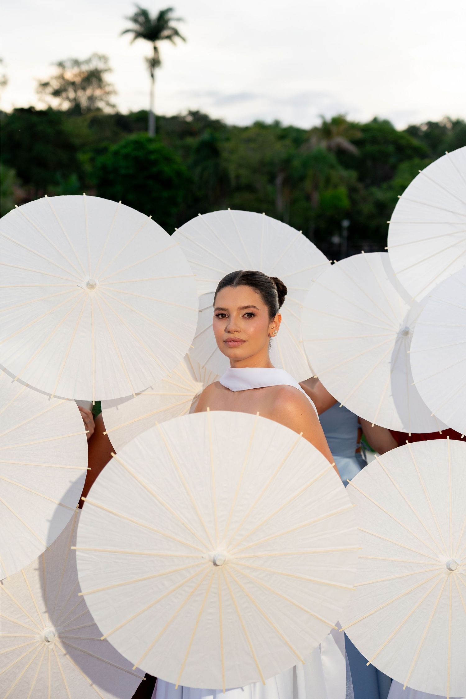 Elegant bride surrounded by white parasols outdoors with lush green trees in the background.