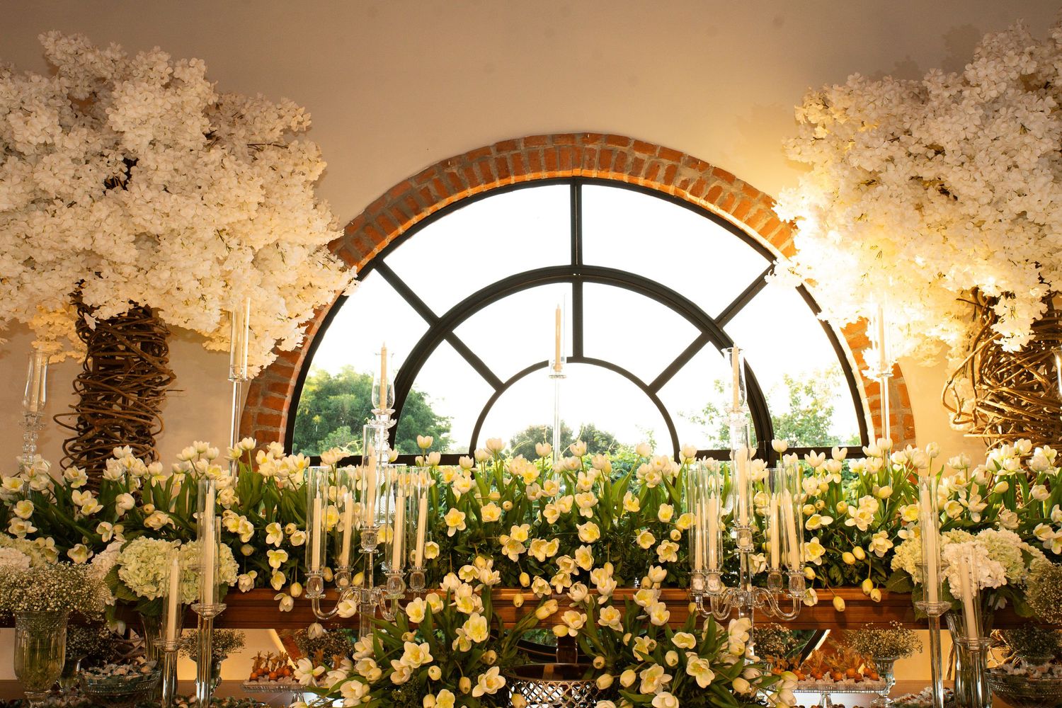 Elegant wedding reception decor with white floral arrangements, candles, and arched window backdrop.