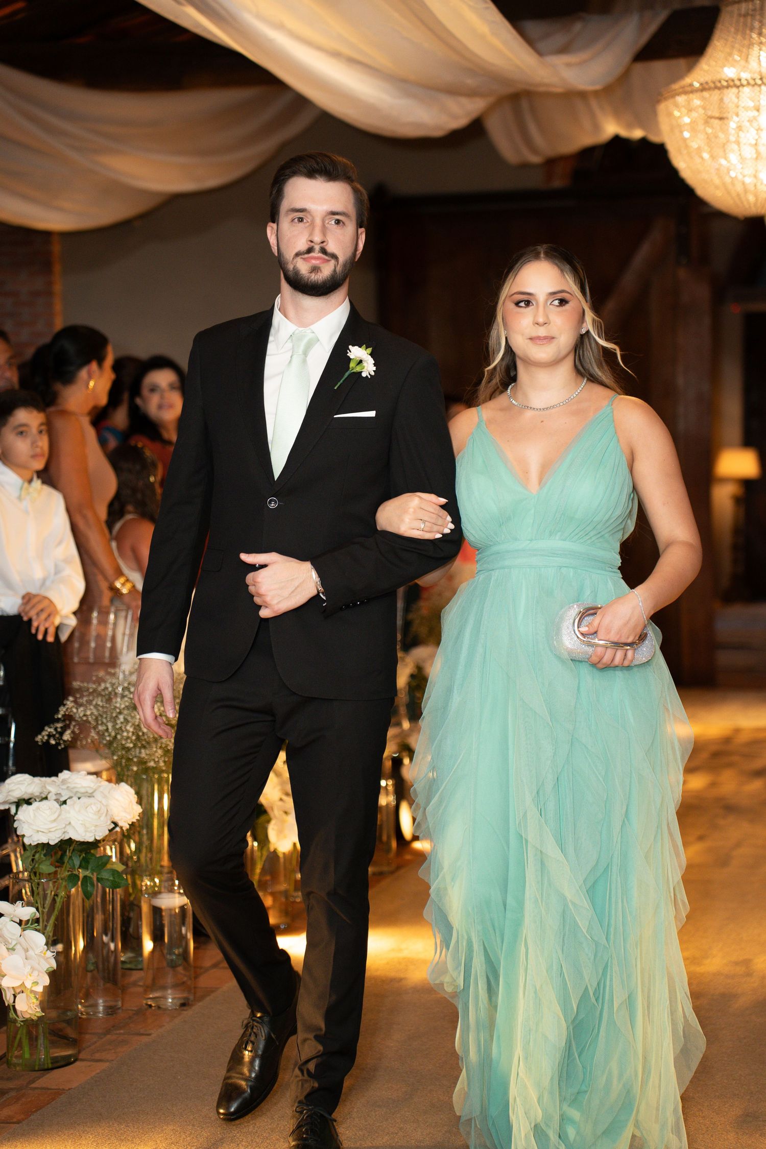 A man in a black tuxedo and a woman in a teal tulle gown walk arm in arm at an elegant wedding reception.
