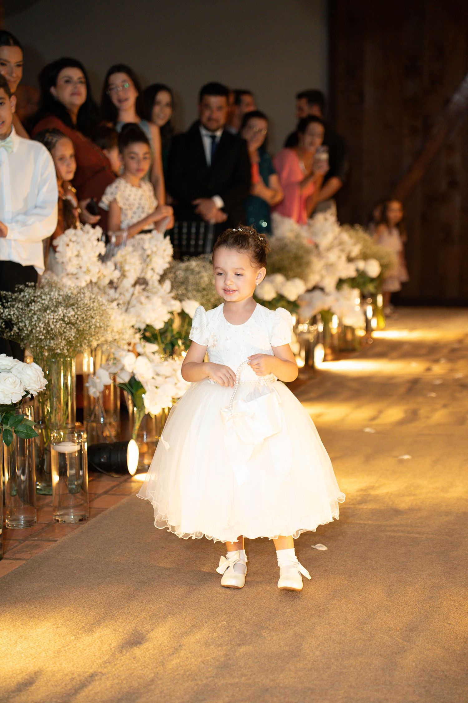 Adorable flower girl in white tulle dress walks down wedding aisle lined with white floral arrangements and candles.