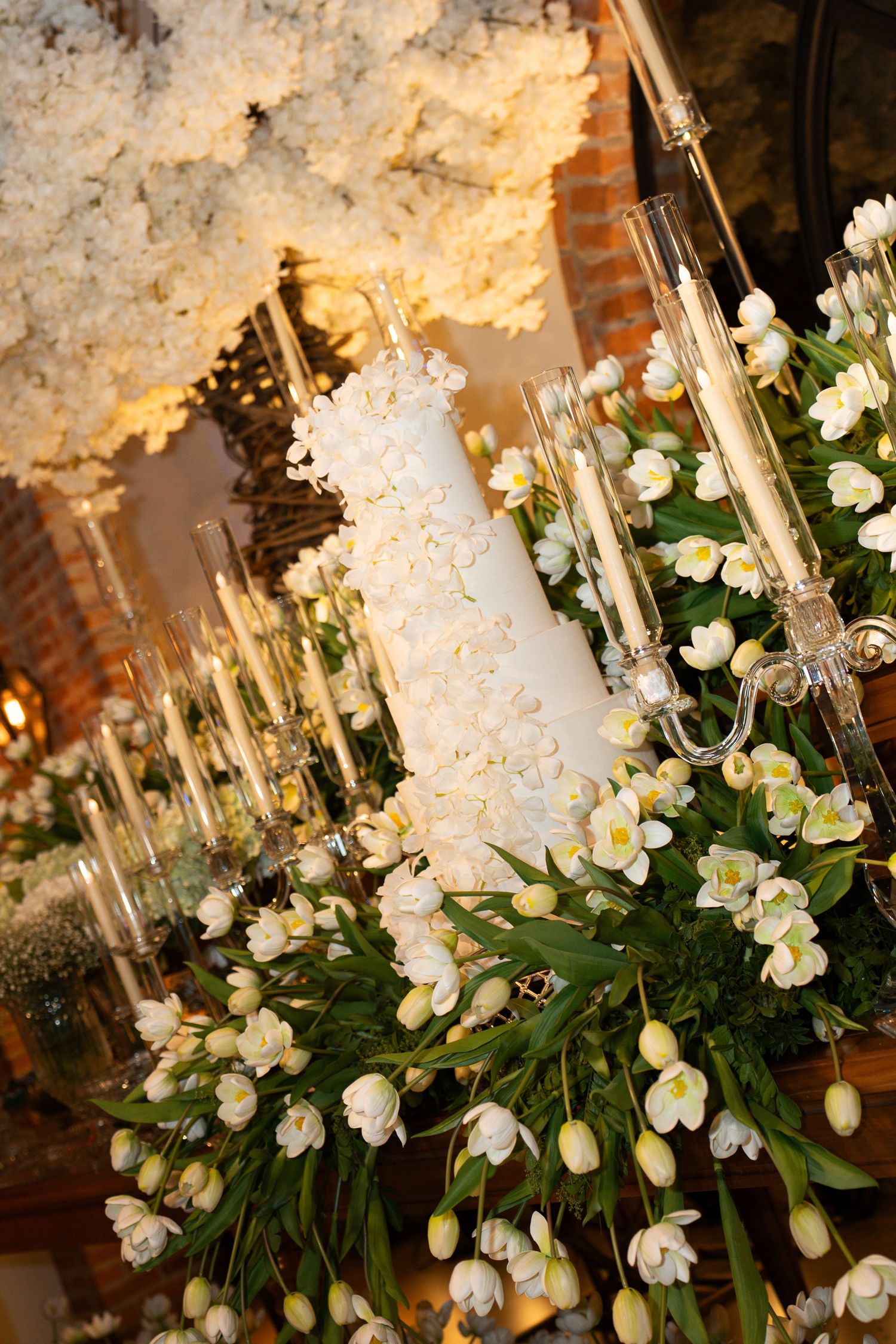 Elegant white wedding cake adorned with flowers, surrounded by white tulips and candelabras in a warmly lit venue.