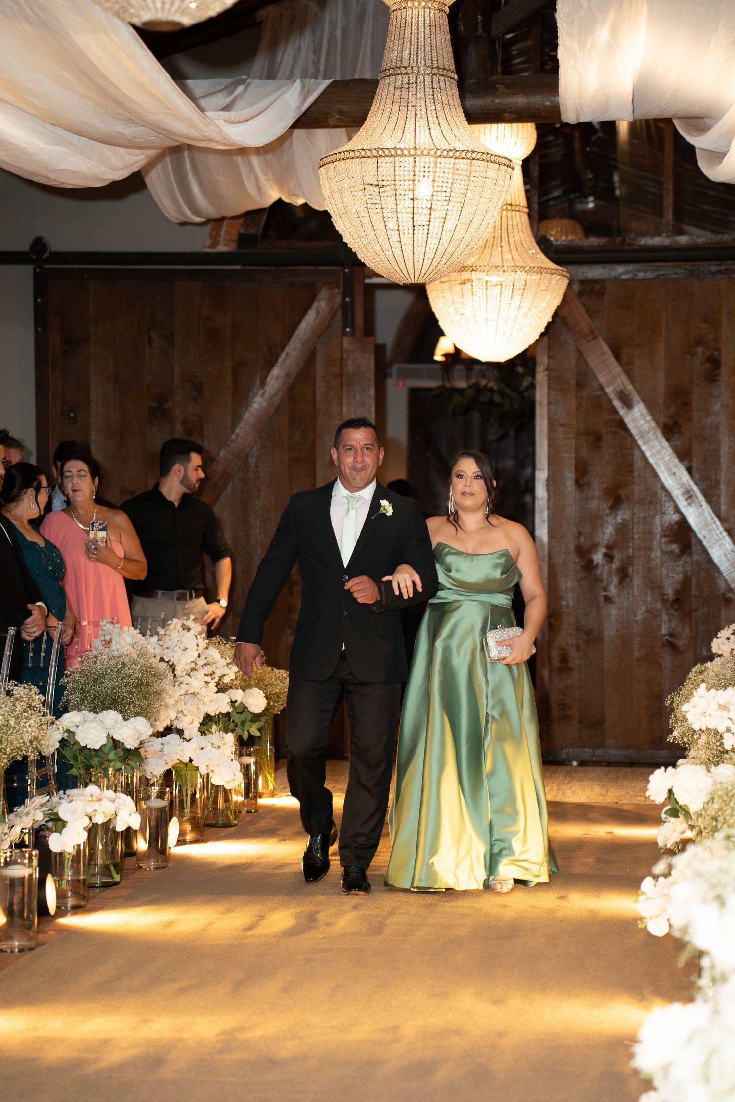 Elegant wedding processional in a barn venue with chandeliers, man in tuxedo escorts woman in green satin gown down floral aisle.