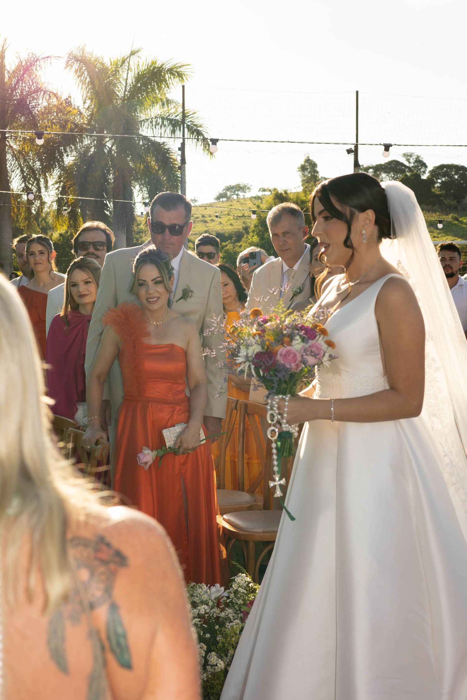 Bride in white gown holding colorful bouquet smiles at outdoor wedding ceremony with guests in vibrant attire.