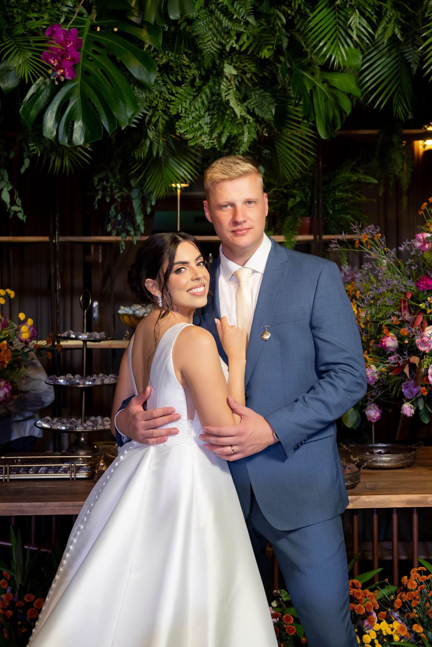 Bride in white gown and groom in blue suit embrace smiling before lush tropical greenery and colorful floral wedding decor.