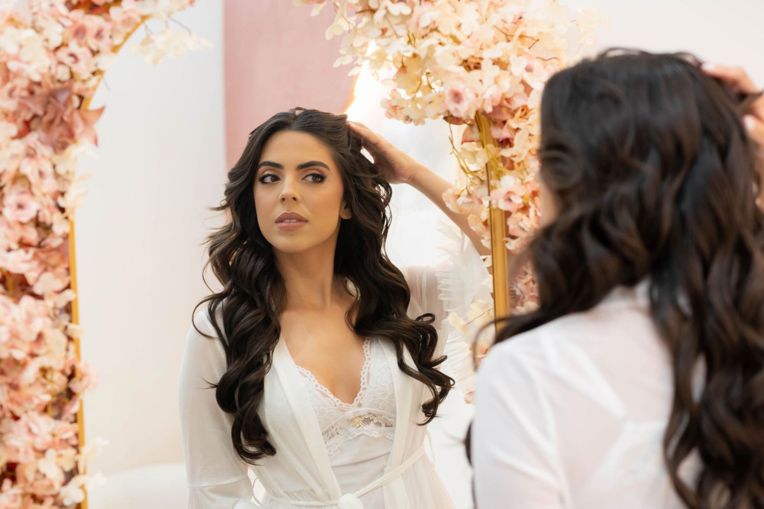 Bride in white robe with long wavy dark hair adjusting her hair in a mirror surrounded by pink floral decor.