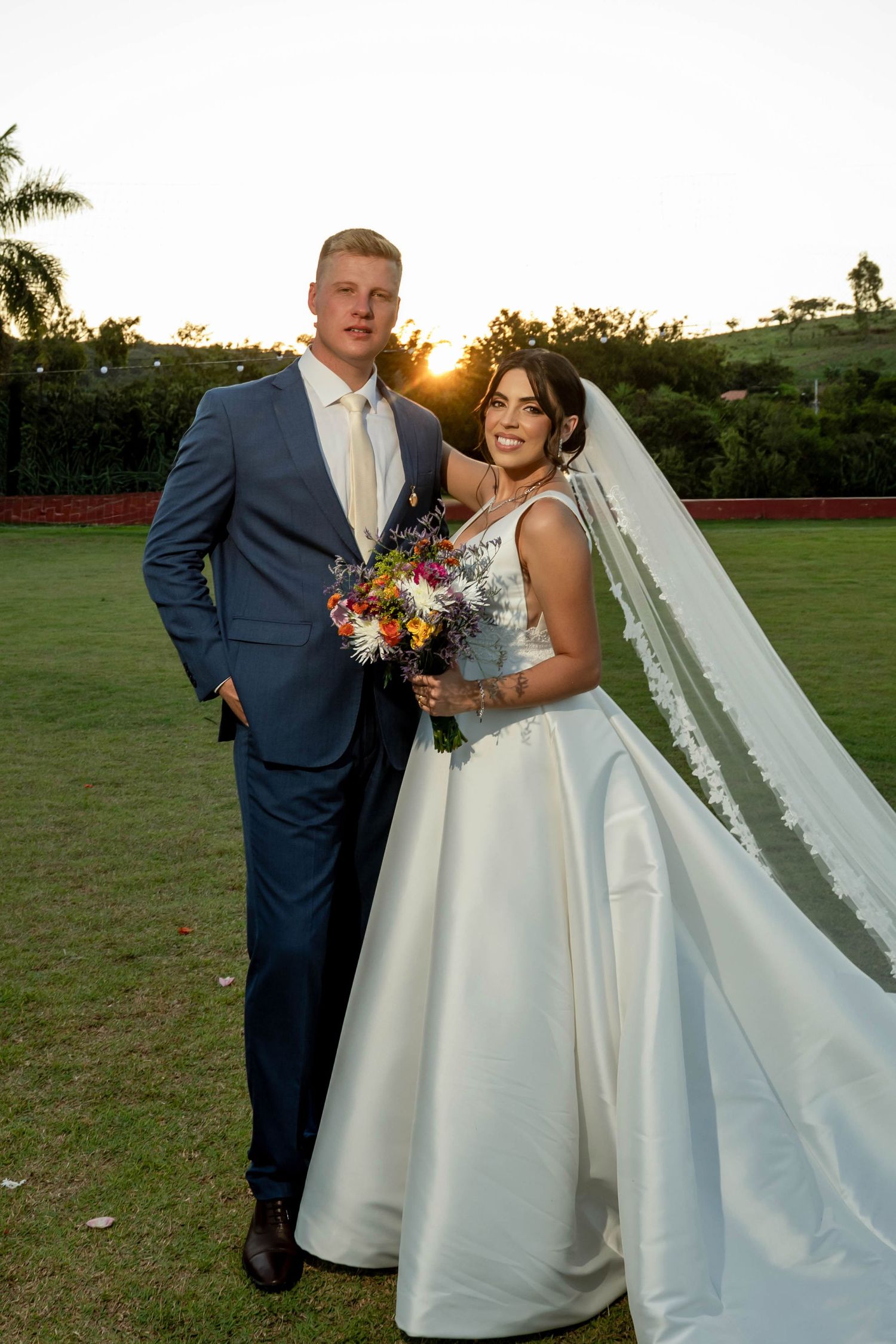 Bride and groom posing at sunset on green lawn, bride in white gown with veil holding colorful bouquet, groom in blue suit.