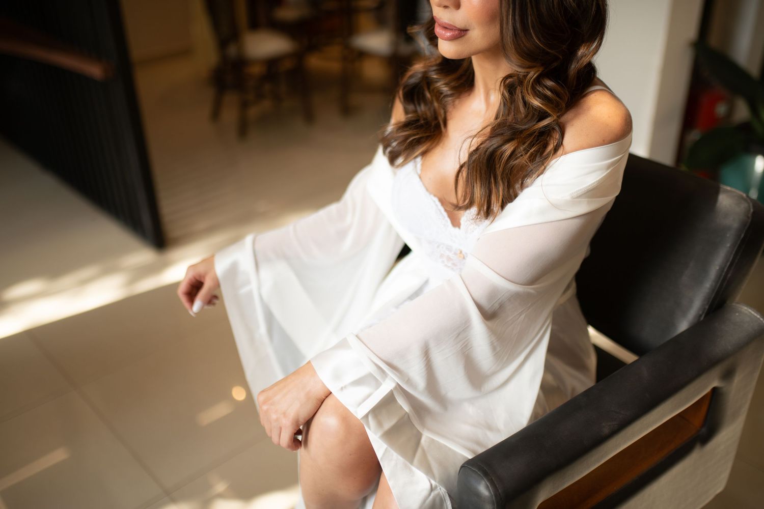 Woman in white off-shoulder robe sitting elegantly in a leather chair, bathed in warm natural sunlight.