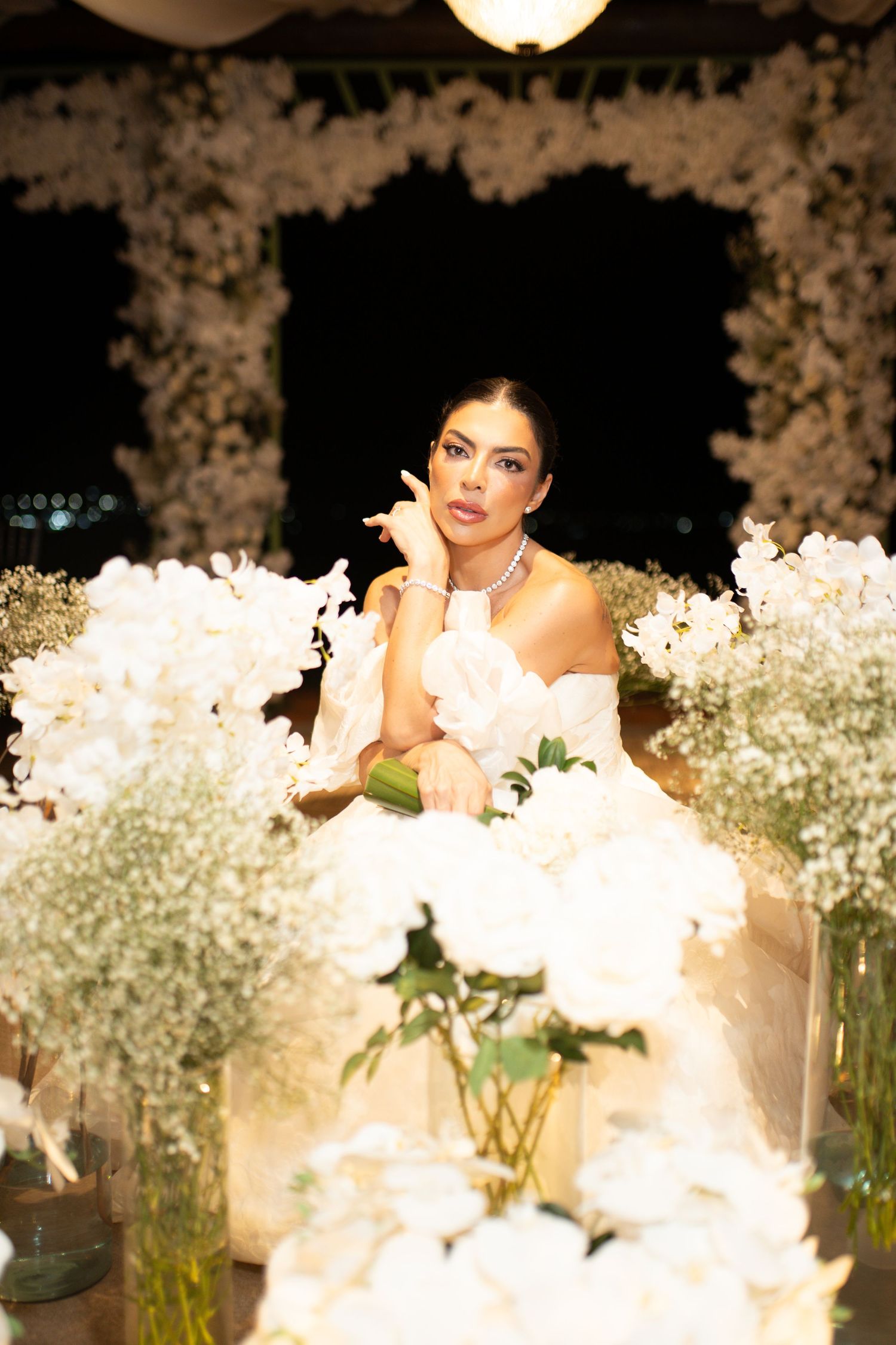 Elegant bride in white off-shoulder gown surrounded by lush white floral arrangements at a luxurious wedding reception.