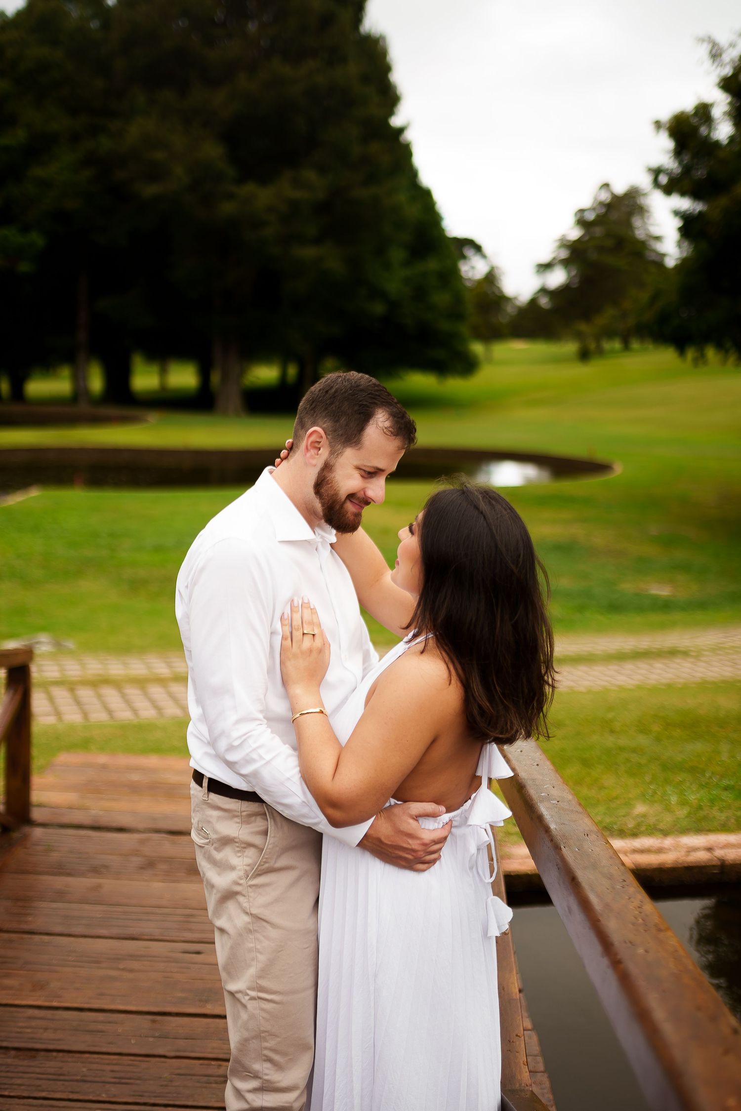 Couple in white embracing on wooden bridge with lush green golf course and pond in background.
