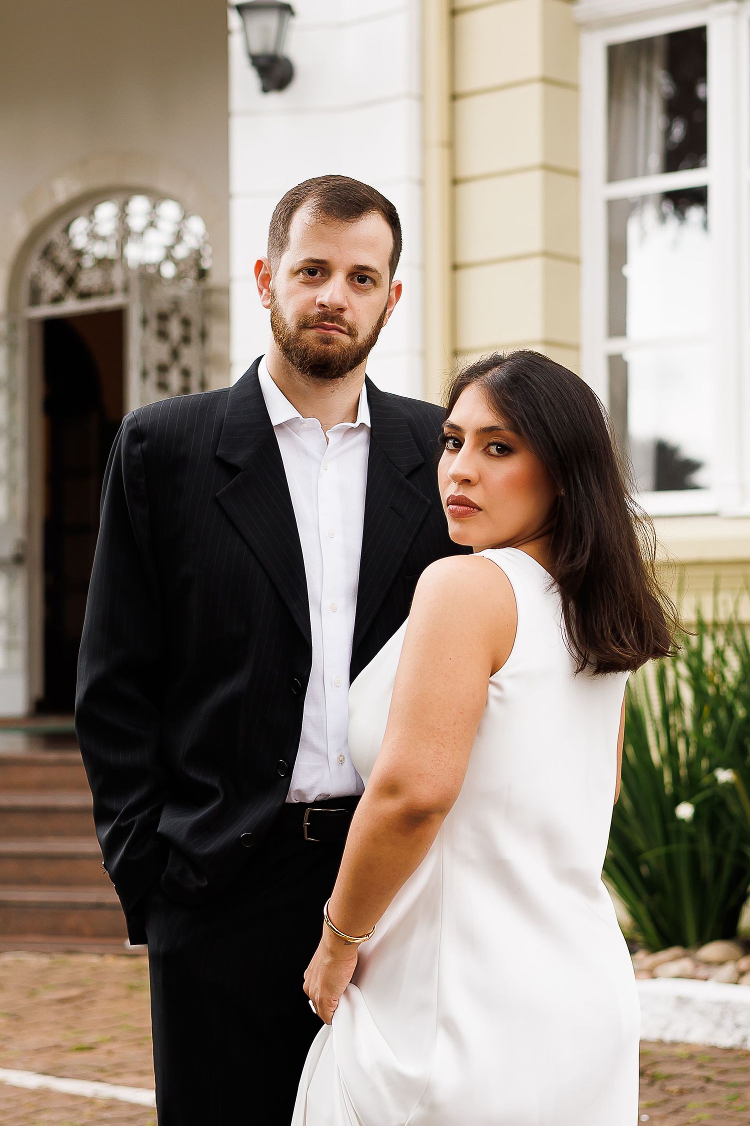 Elegant couple posing in formal attire, man in black suit, woman in white dress, standing before a classic building.