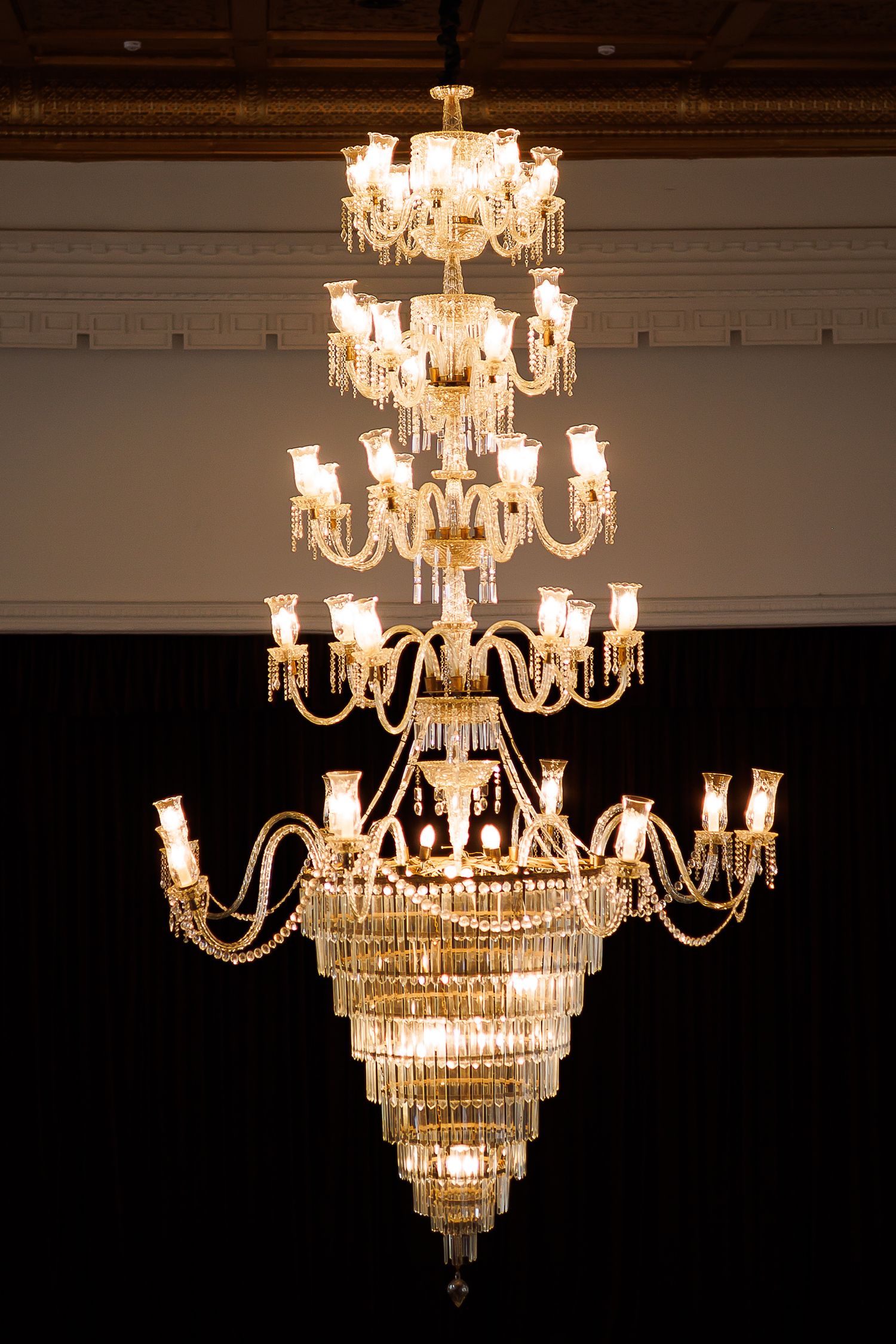 Elegant multi-tiered crystal chandelier with gold accents and glowing candle lights hanging in a grand ballroom.