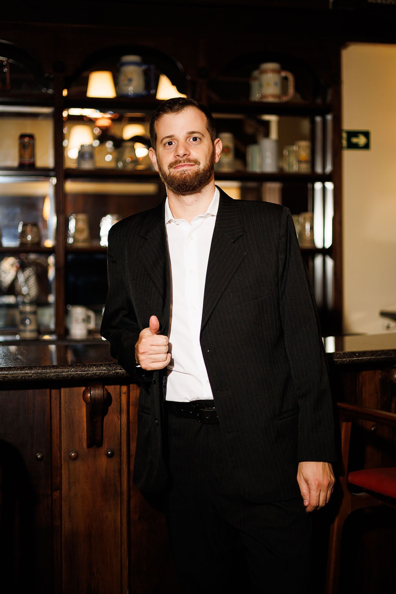 Bearded man in black suit giving thumbs up while standing at a bar counter in a classic restaurant.