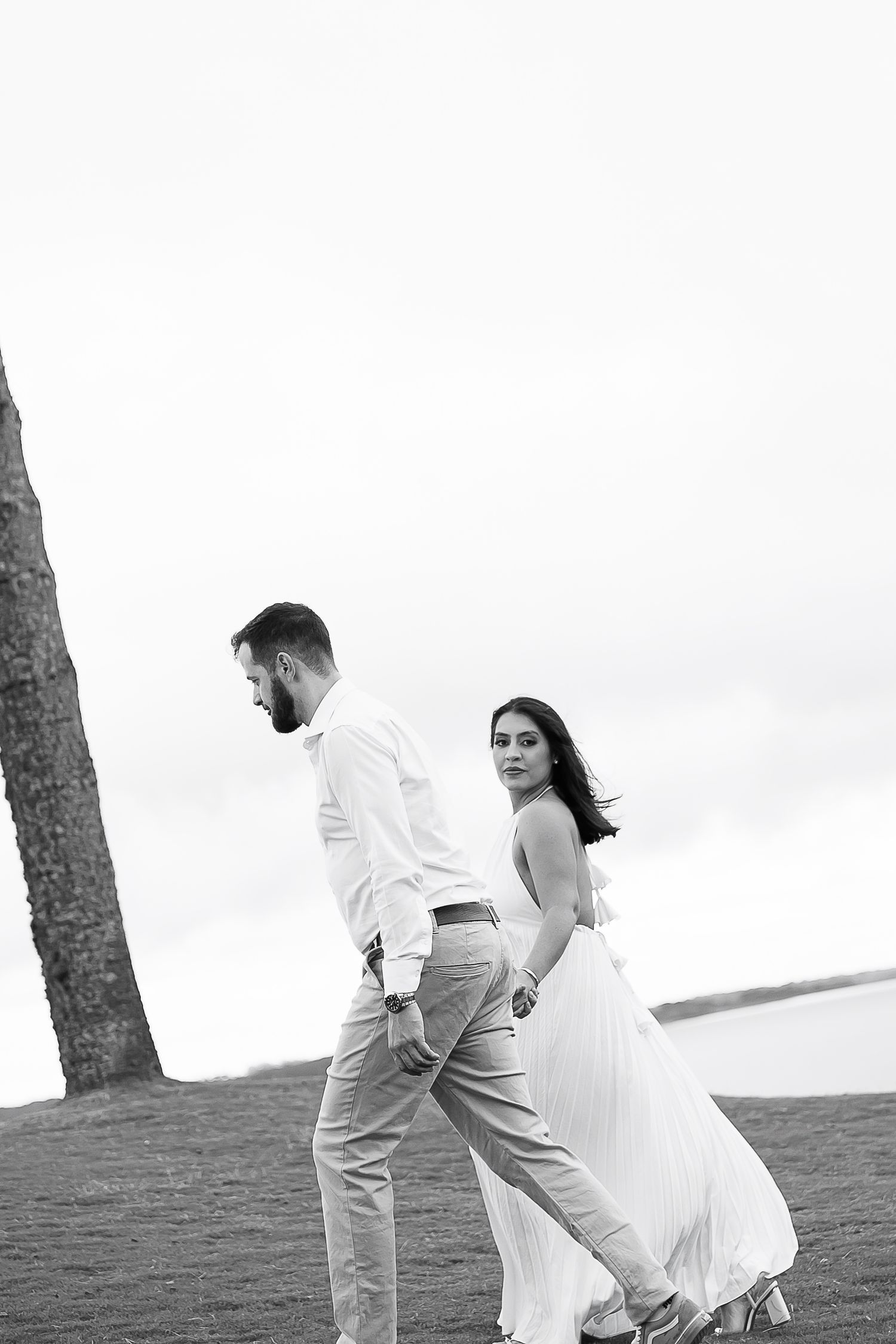 Black and white photo of an elegant couple walking hand in hand near the ocean, woman in flowing white dress.